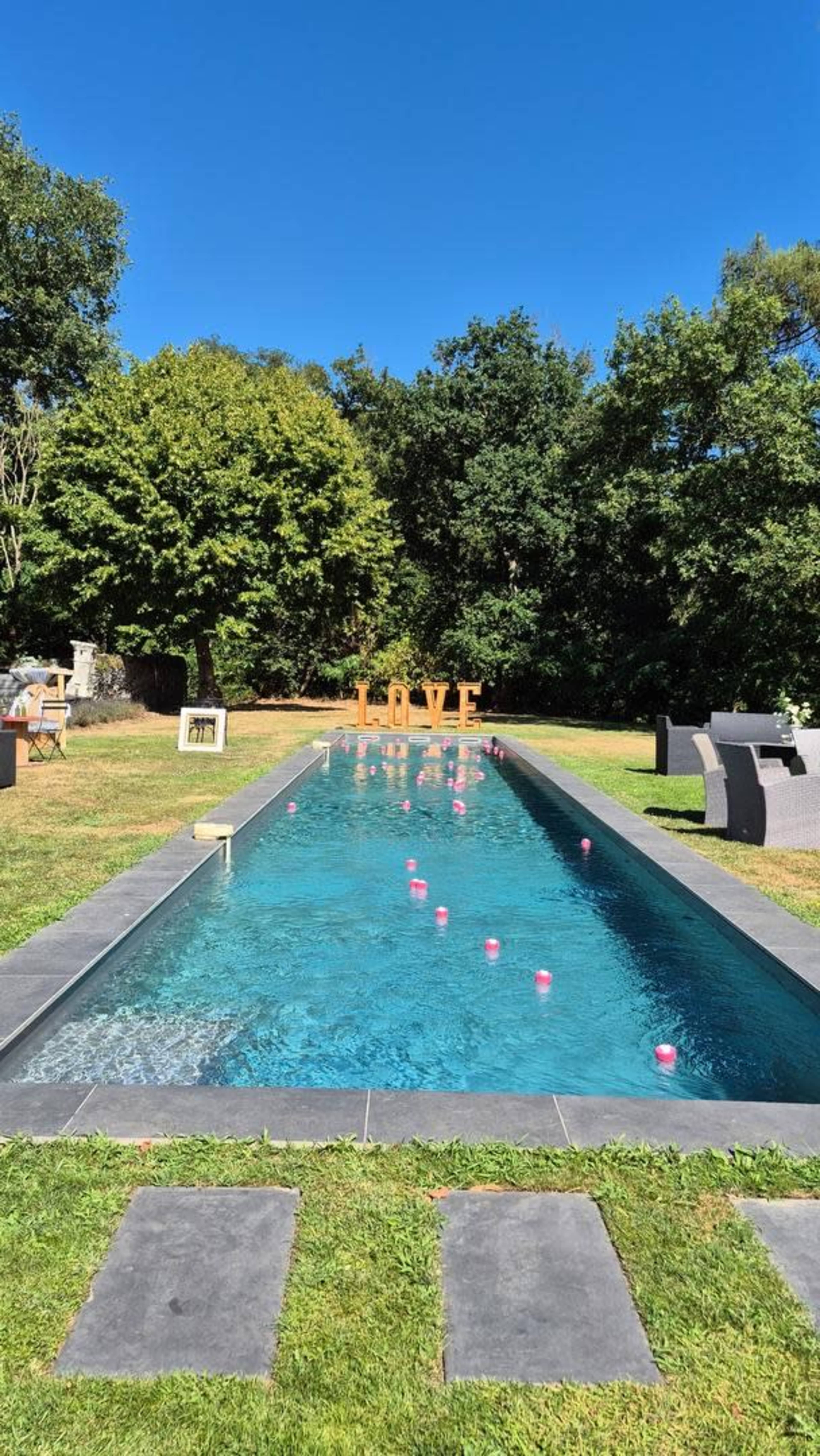 The image shows a rectangular swimming pool in a grassy area, with pink floats scattered on the water and large "LOVE" letters on the opposite side against a backdrop of trees.