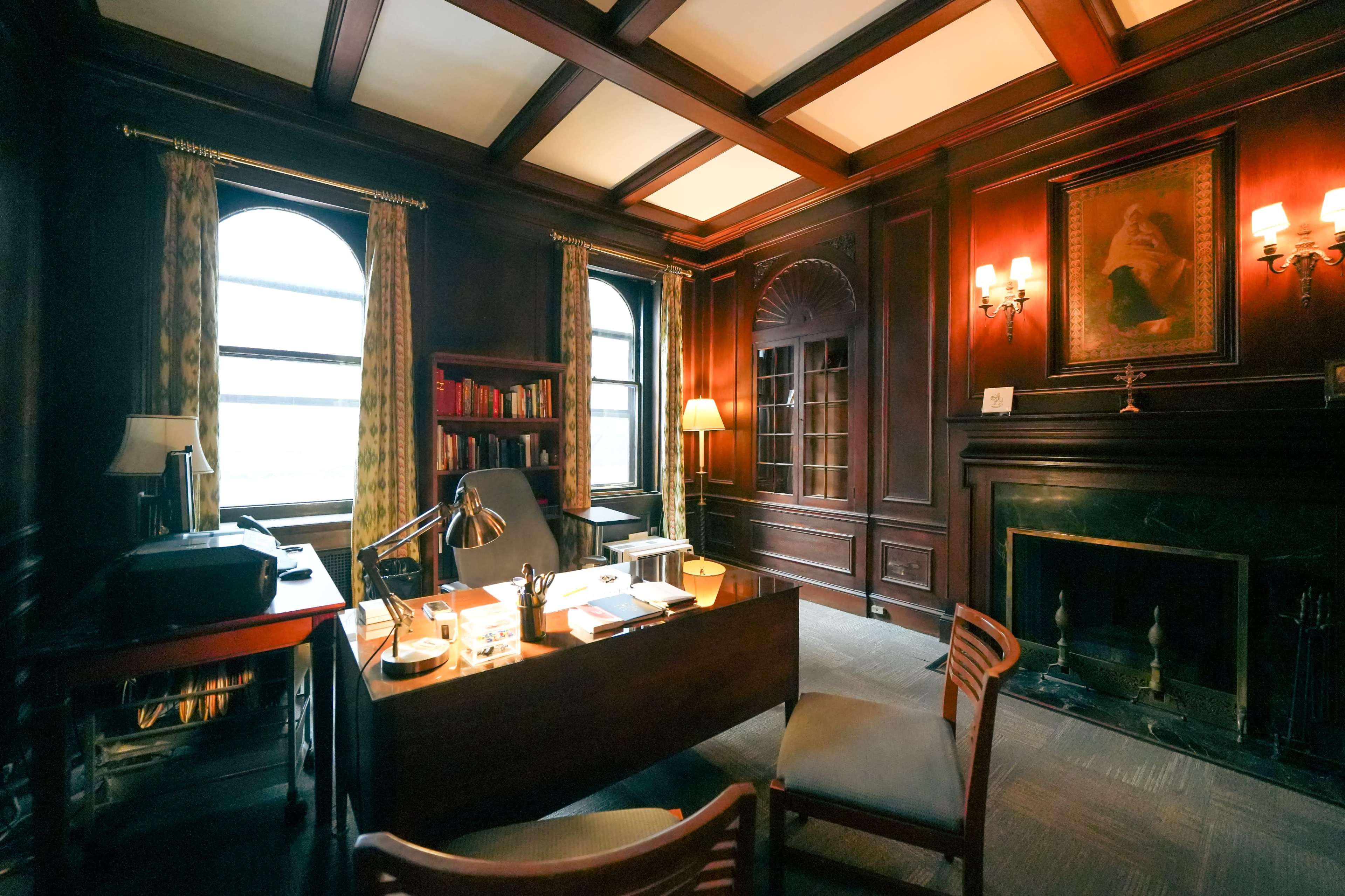 The image shows a dark wood-paneled office with a large desk, bookshelves, and two windows.