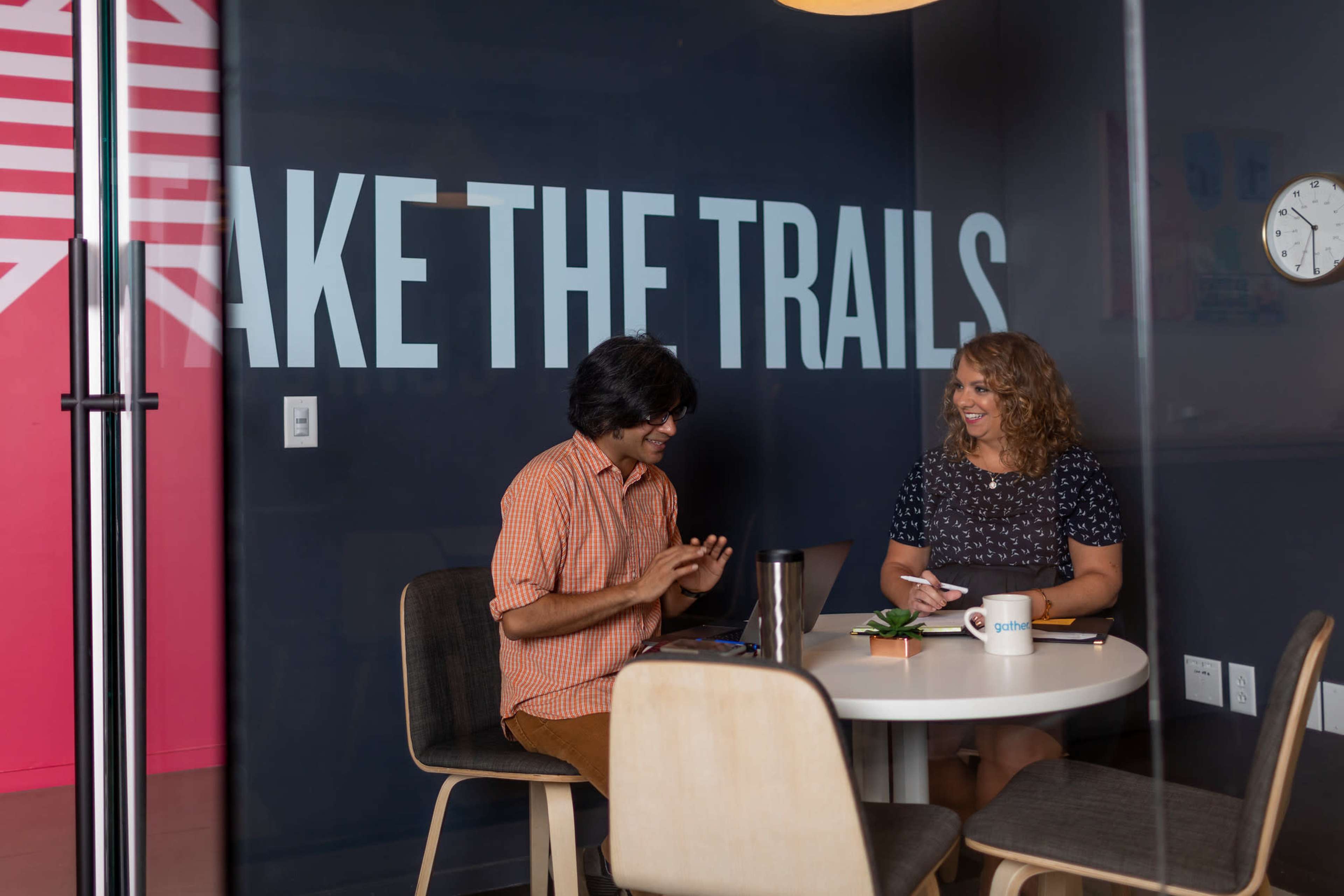 Two individuals are seated at a round table in a meeting room, engaged in discussion, with a large sign on the wall that reads "MAKE THE TRAILS."
