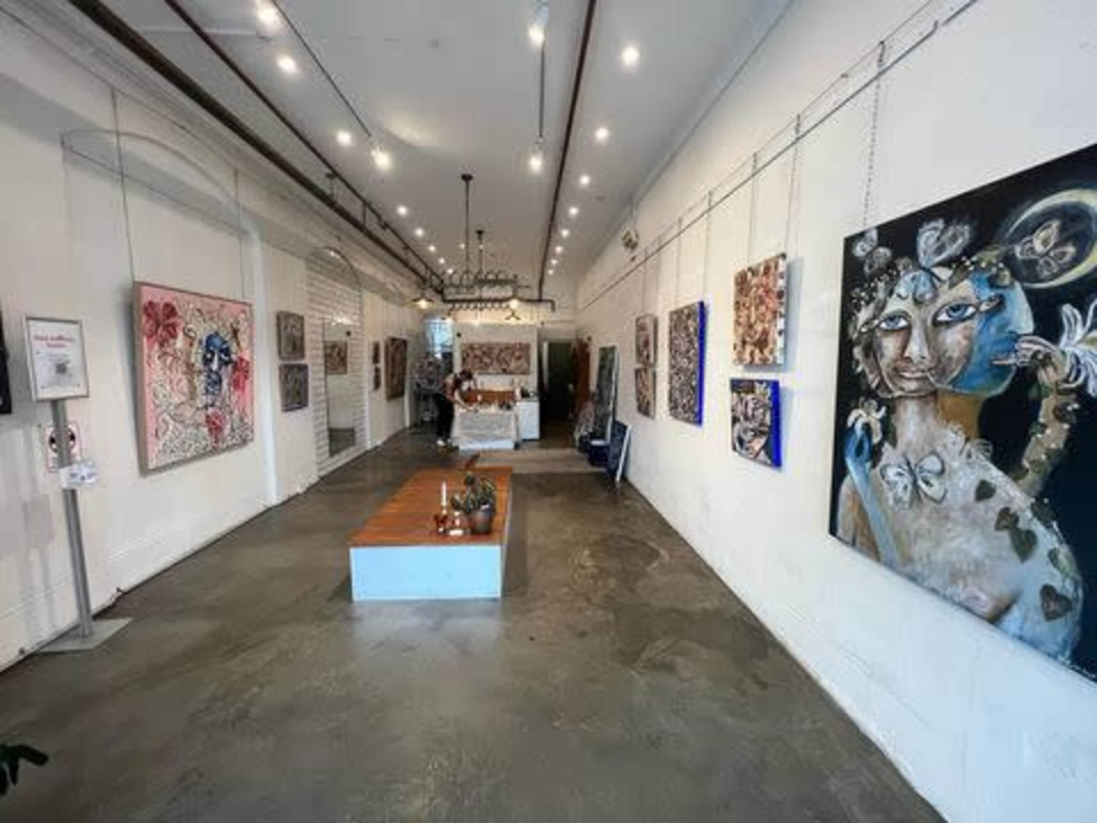 A modern art gallery with paintings displayed on the white walls and a wooden table in the center of the space.