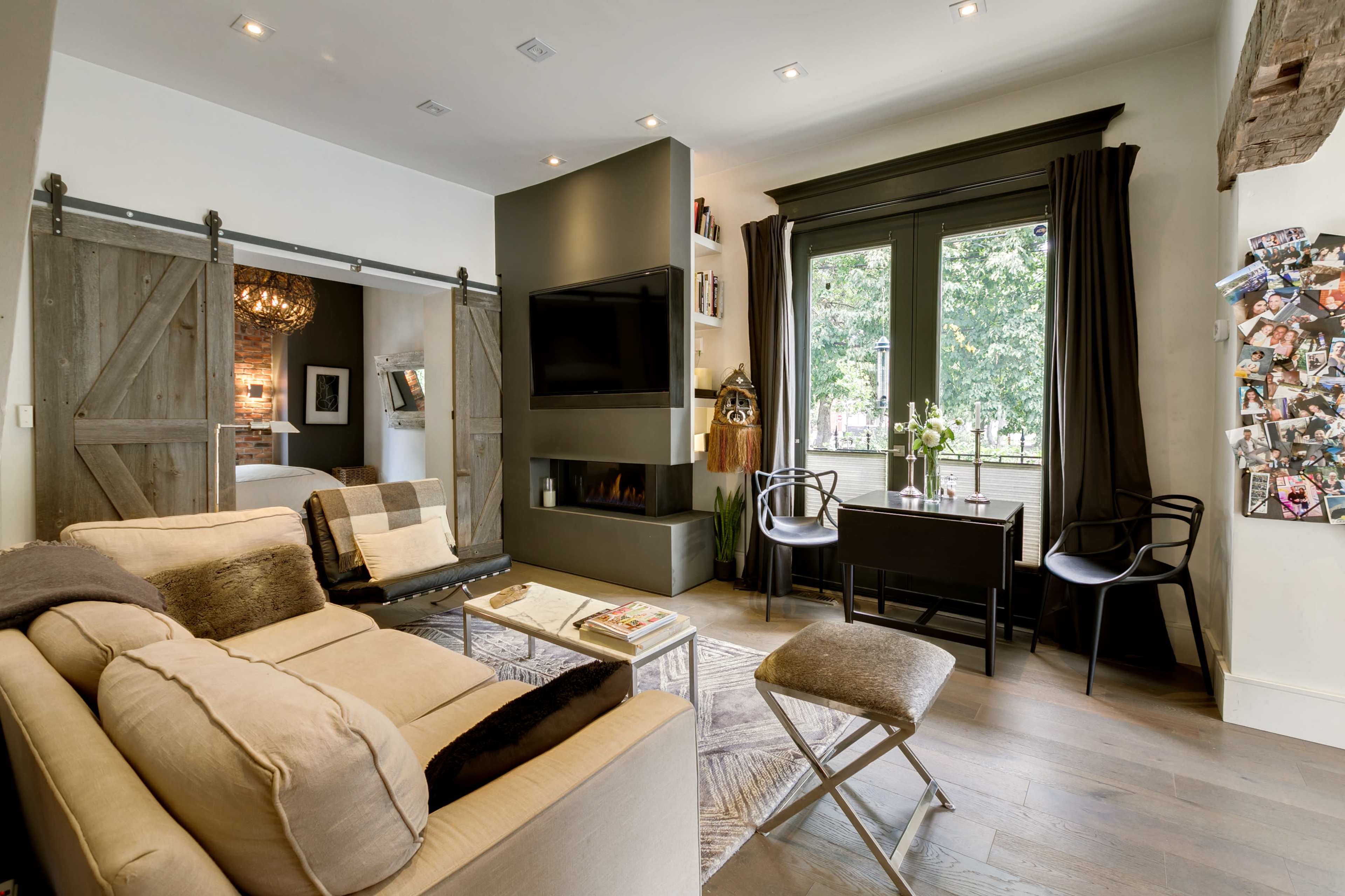 A modern living room with a sofa, a coffee table, and a dining area by a window, featuring a sliding barn door and a wall-mounted fireplace.
