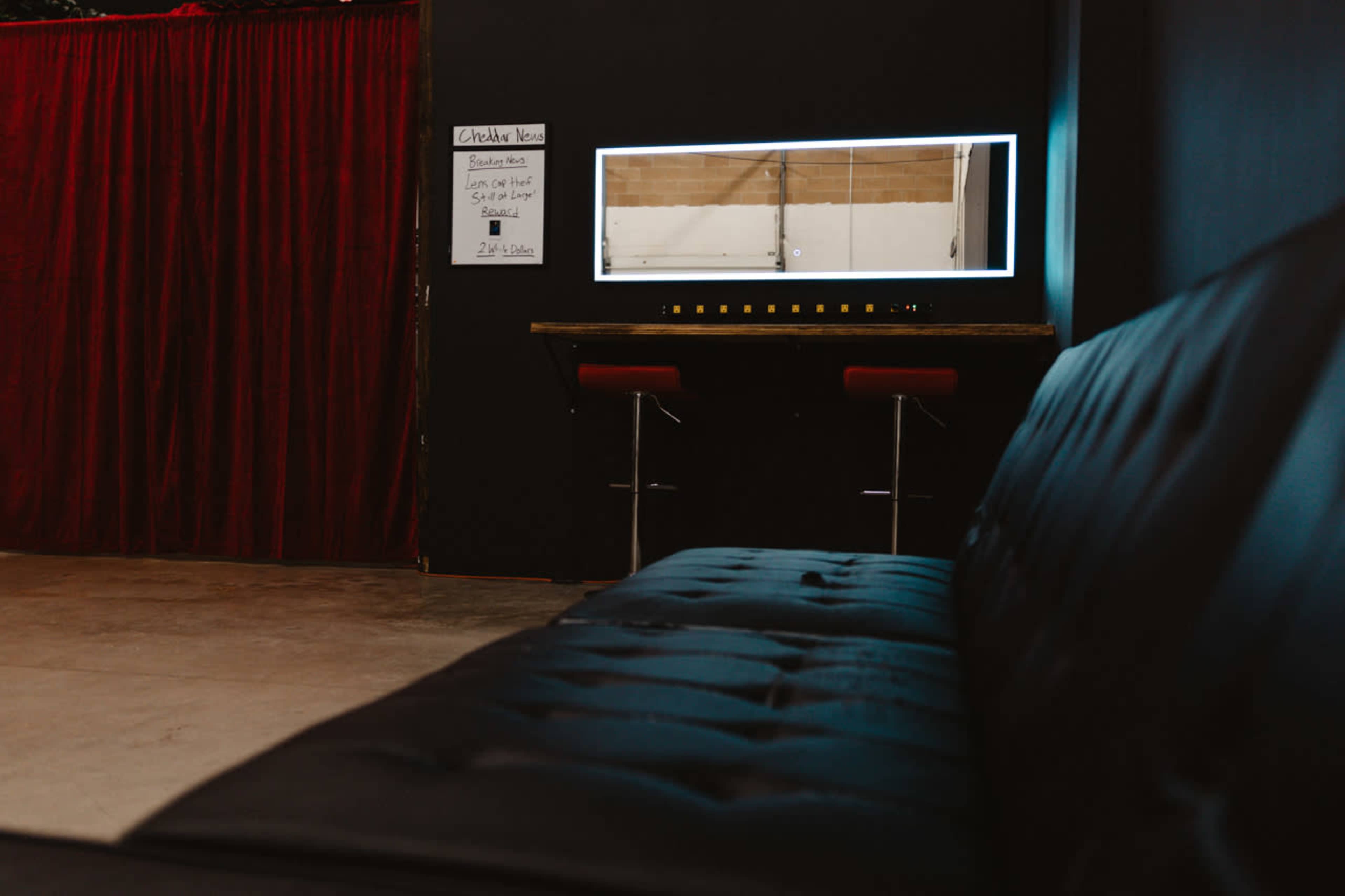A black couch is positioned in front of a brightly lit mirror against a dark wall, with a curtain and a sign listing services visible in the background.