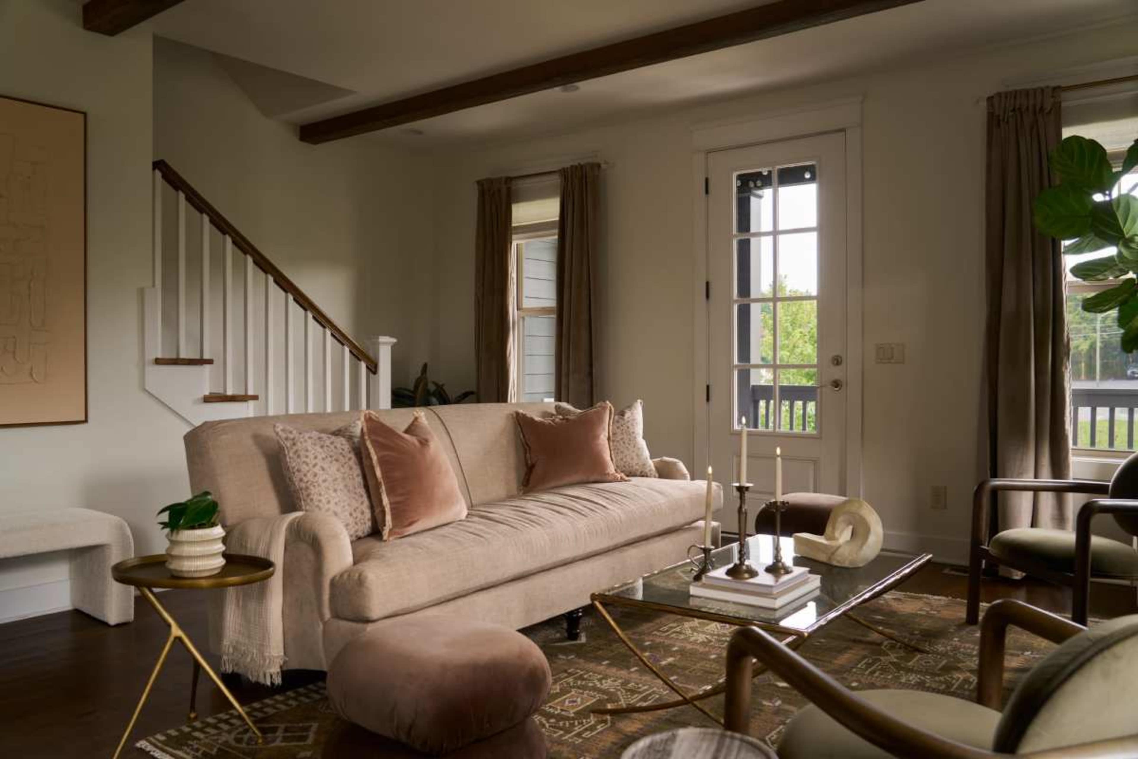 The image shows a cozy living room with a beige sofa, a decorative rug, and a glass coffee table, featuring large windows with natural light and a staircase in the background.