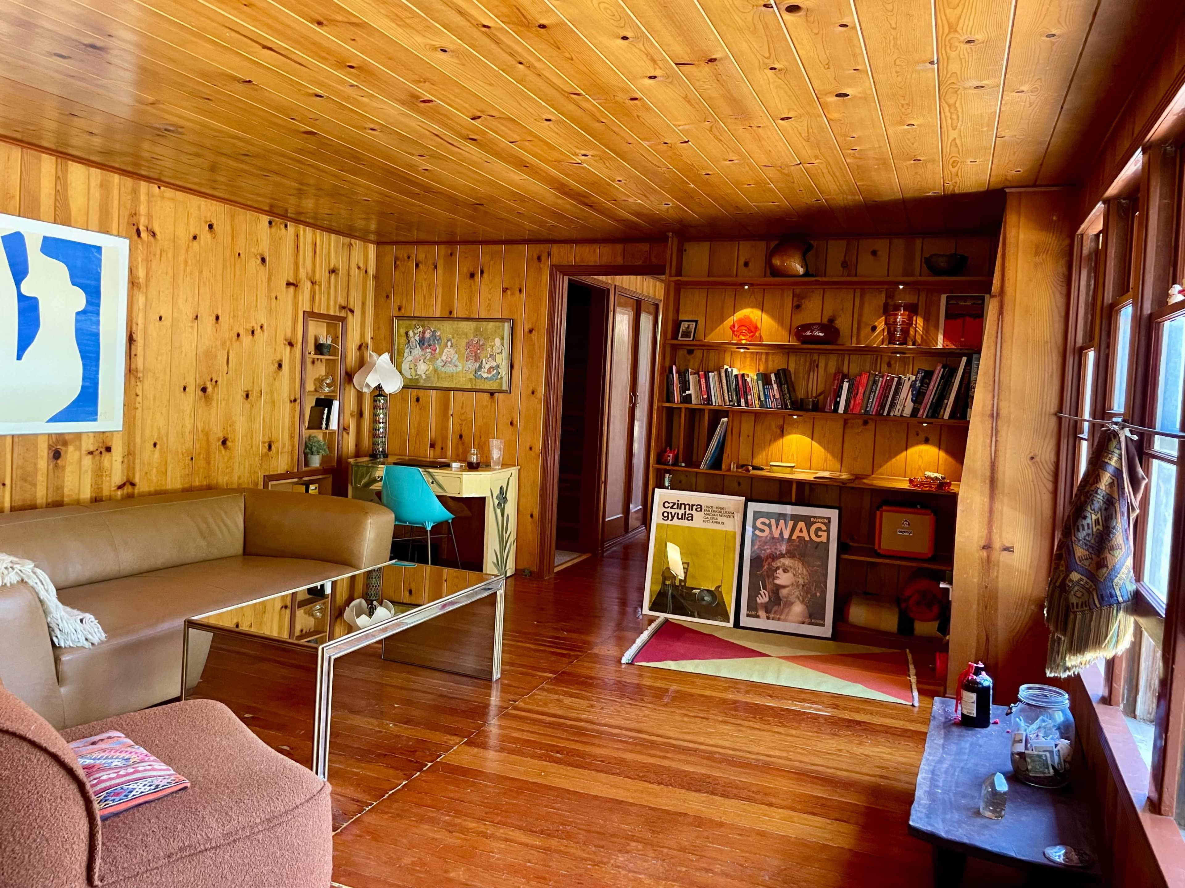 The image shows a wooden-paneled living room with a sofa, a glass coffee table, a bookshelf filled with books, and artwork on the walls.