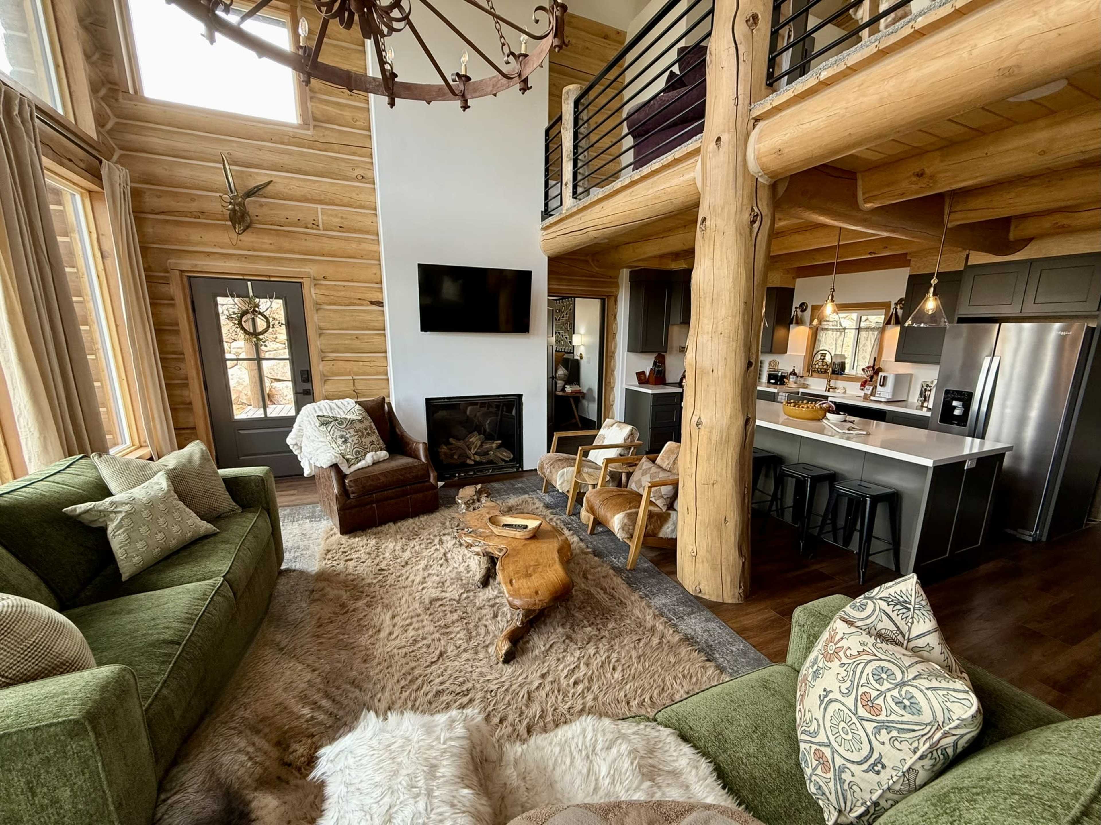 The image shows a cozy living room with a large fireplace, green sofas, and a view of a kitchen area featuring modern appliances and wooden accents.