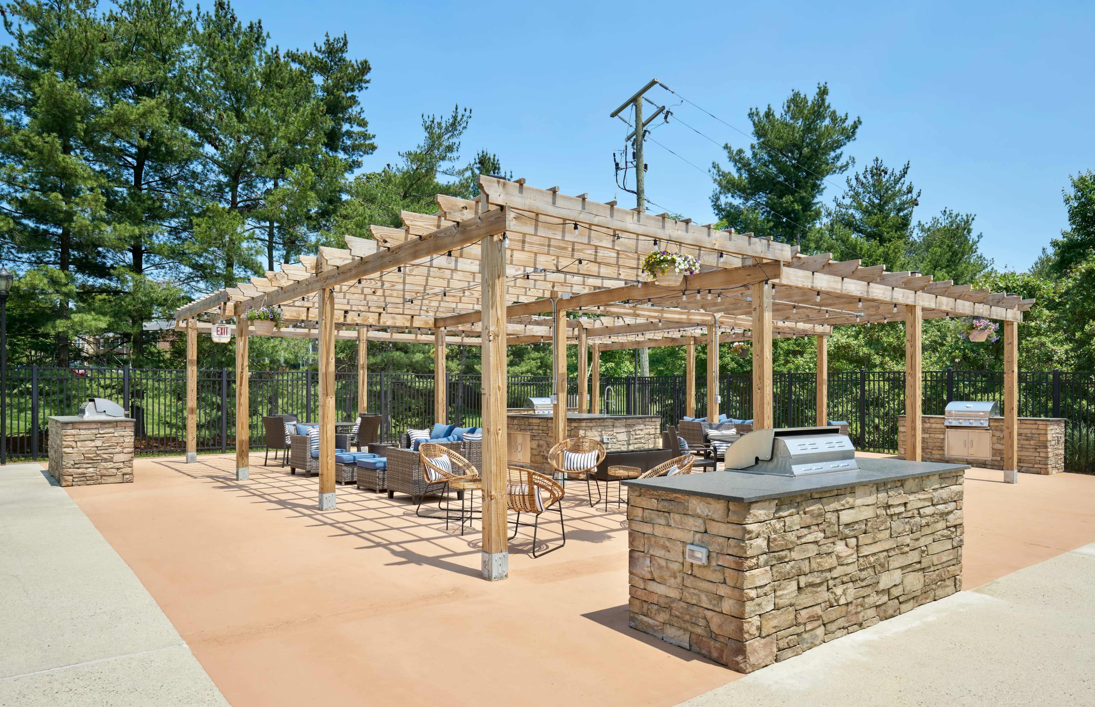 The image shows a spacious outdoor area with a pergola, featuring stone grill stations, seating arrangements, and surrounded by trees.