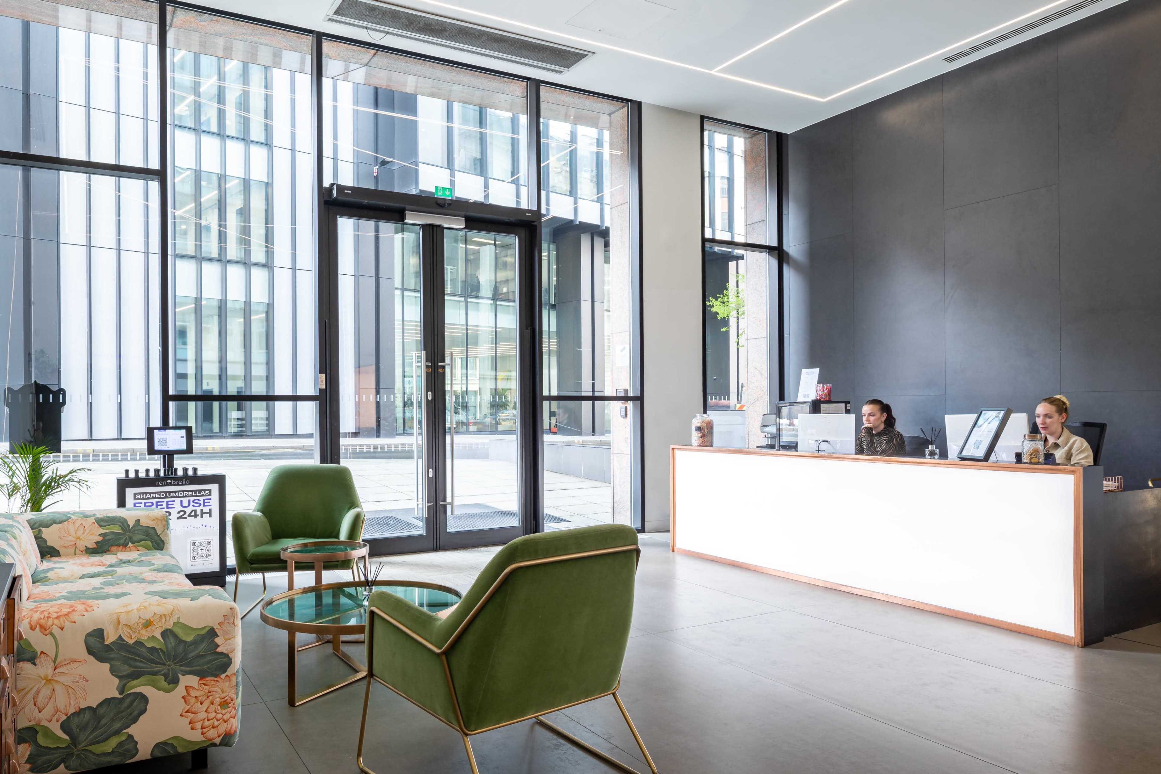 An indoor reception area features a modern desk, two employees, and seating arrangements with greenery visible through large glass doors.