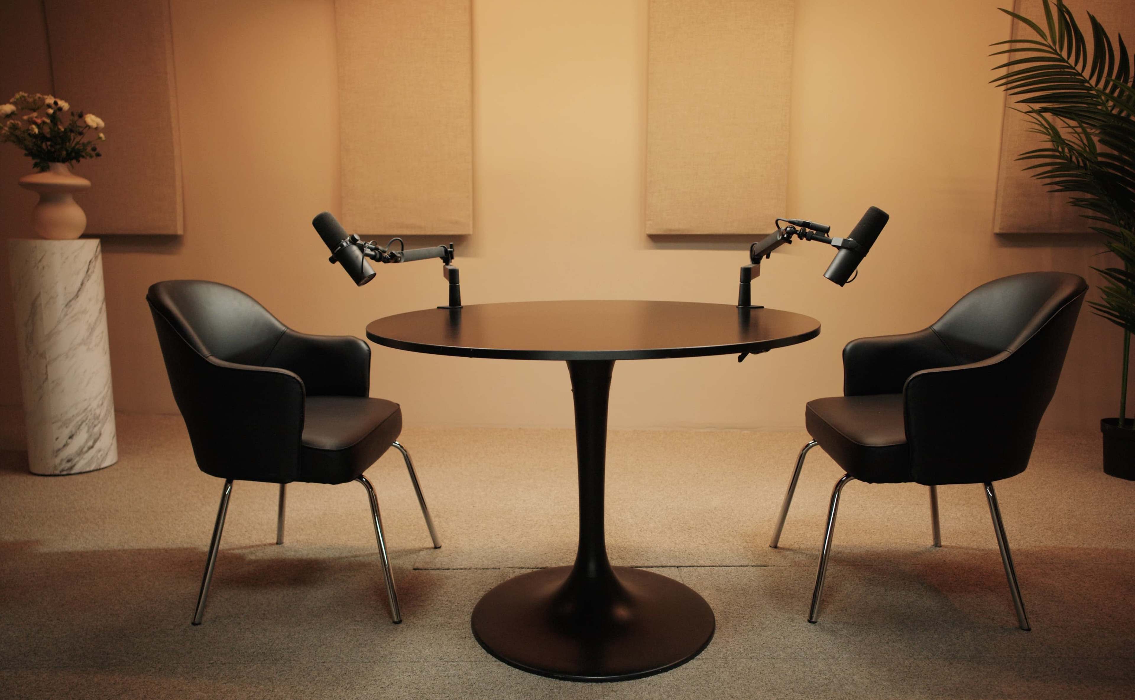 A round black table with two black chairs and mounted microphones is set up in a softly lit room with soundproofing panels.