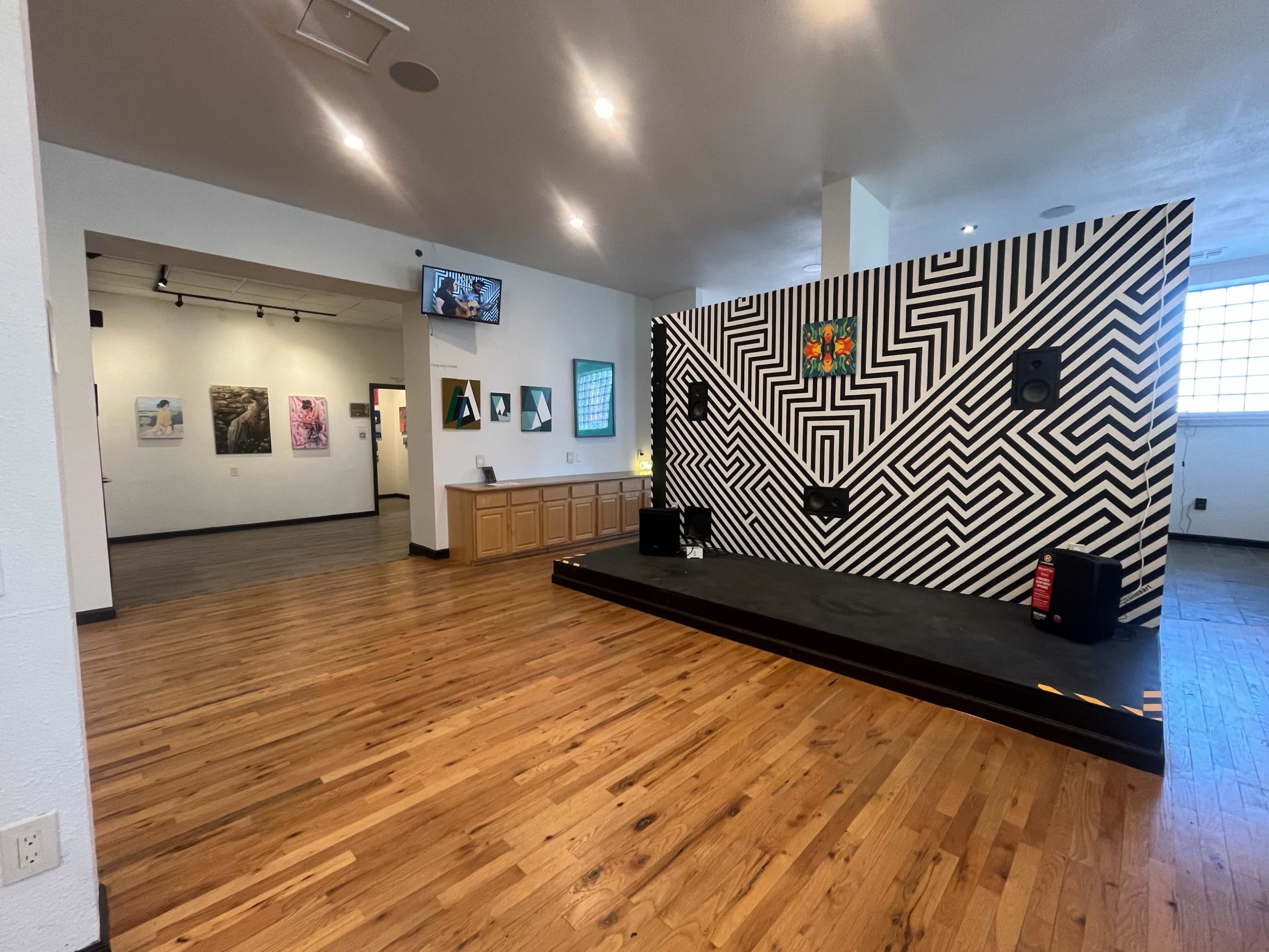 The image shows a modern gallery space with a wooden floor, featuring a striped mural on the wall, a small stage area with speakers, and several framed artworks on display.