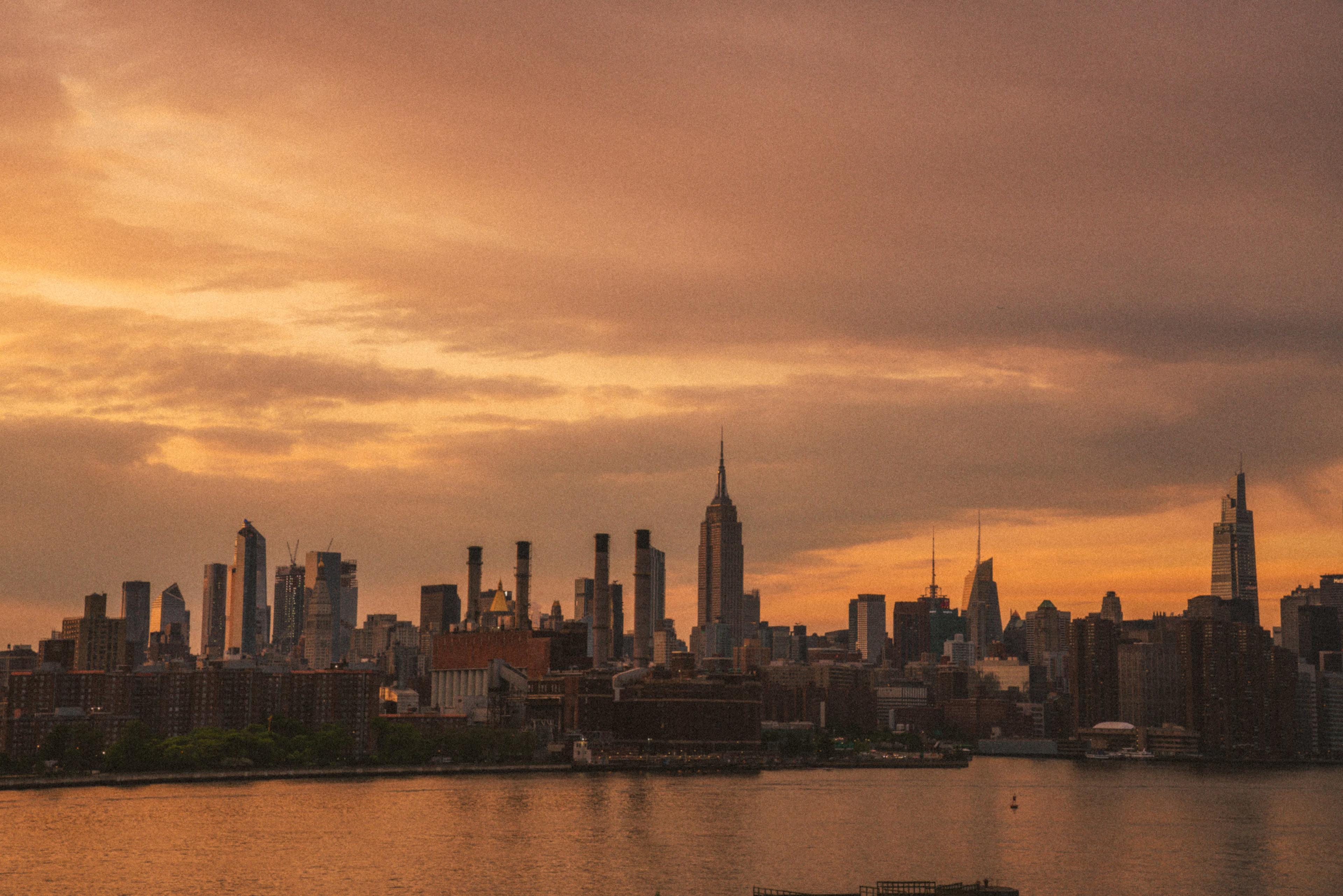 The skyline of New York City is silhouetted against a colorful sunset, featuring notable buildings like the Empire State Building.