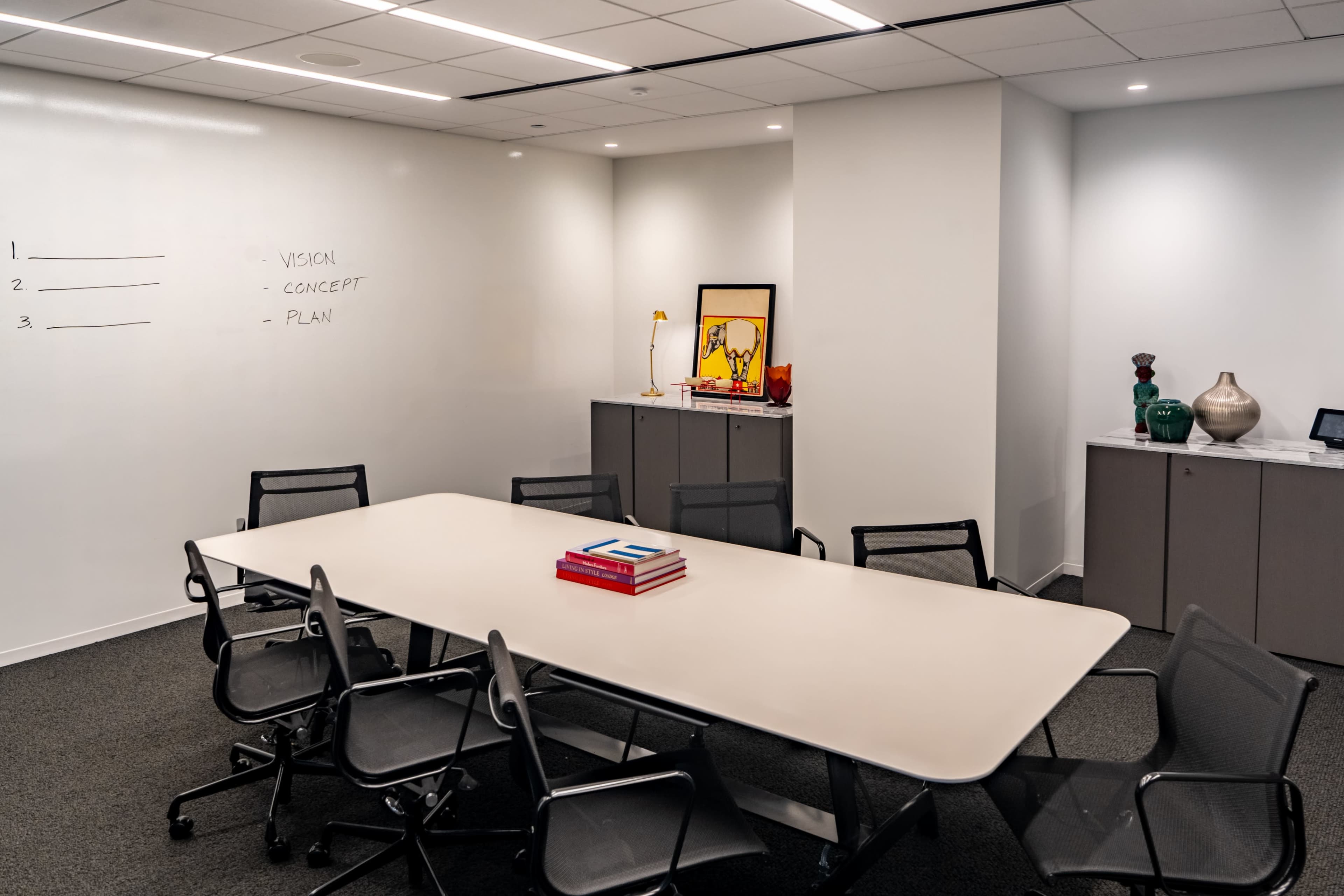 A conference room features a large table surrounded by chairs, with a whiteboard on one wall and a cabinet displaying decorative items on another.