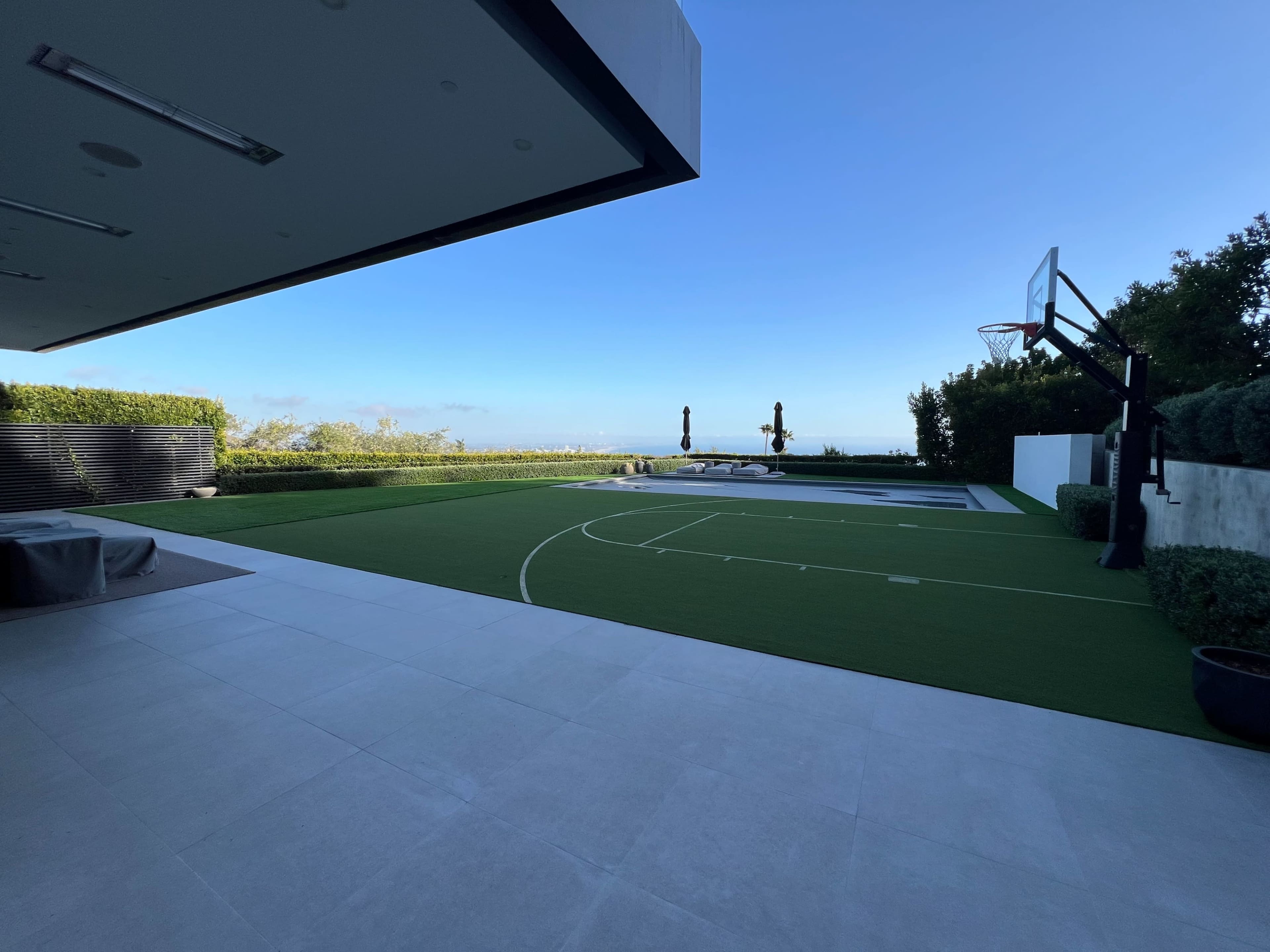 A spacious outdoor area features a basketball court, green turf, and a view of the horizon, with trees lining the perimeter.