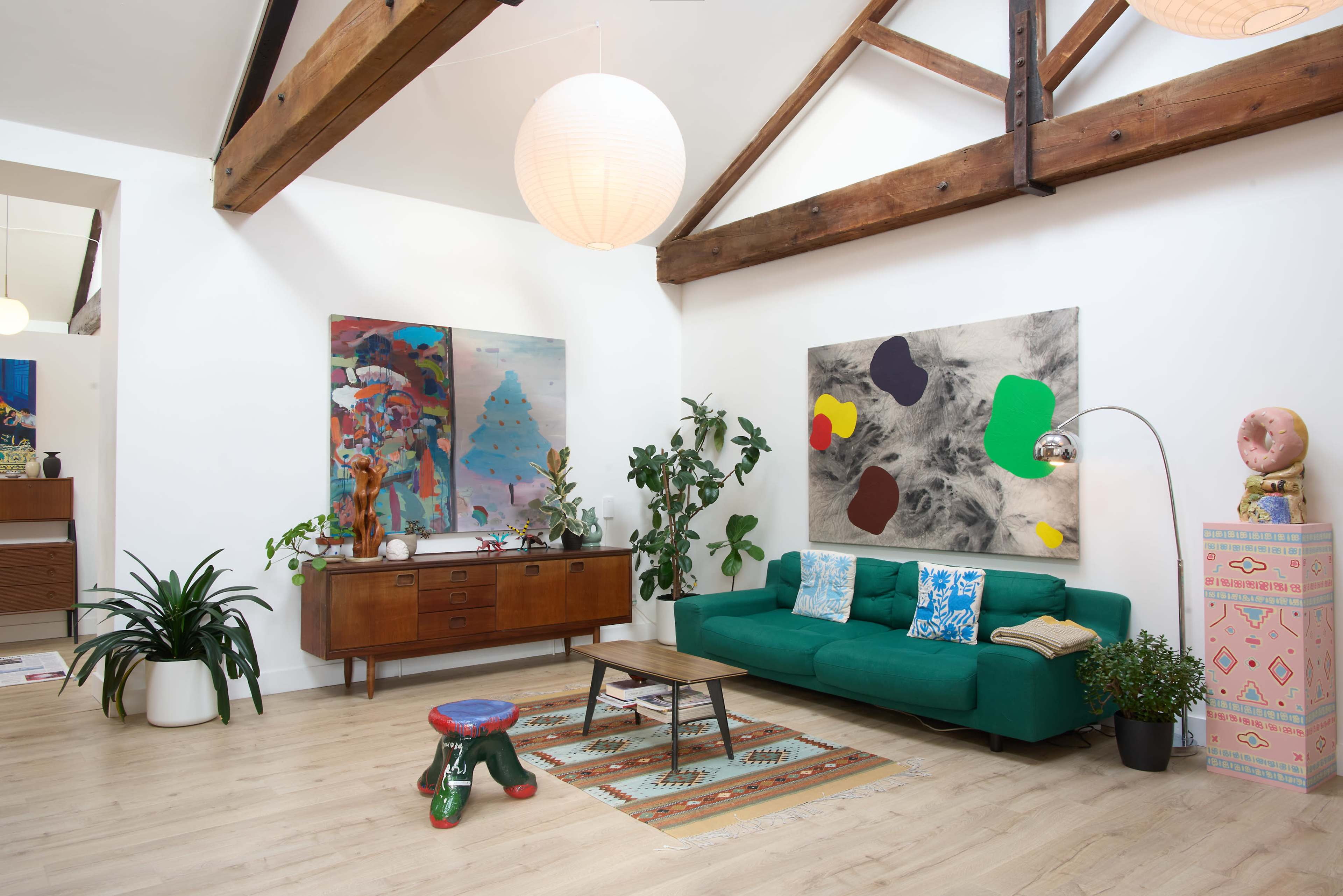 The image shows a modern living room with a green sofa, wooden furniture, colorful artwork on the walls, and various indoor plants.