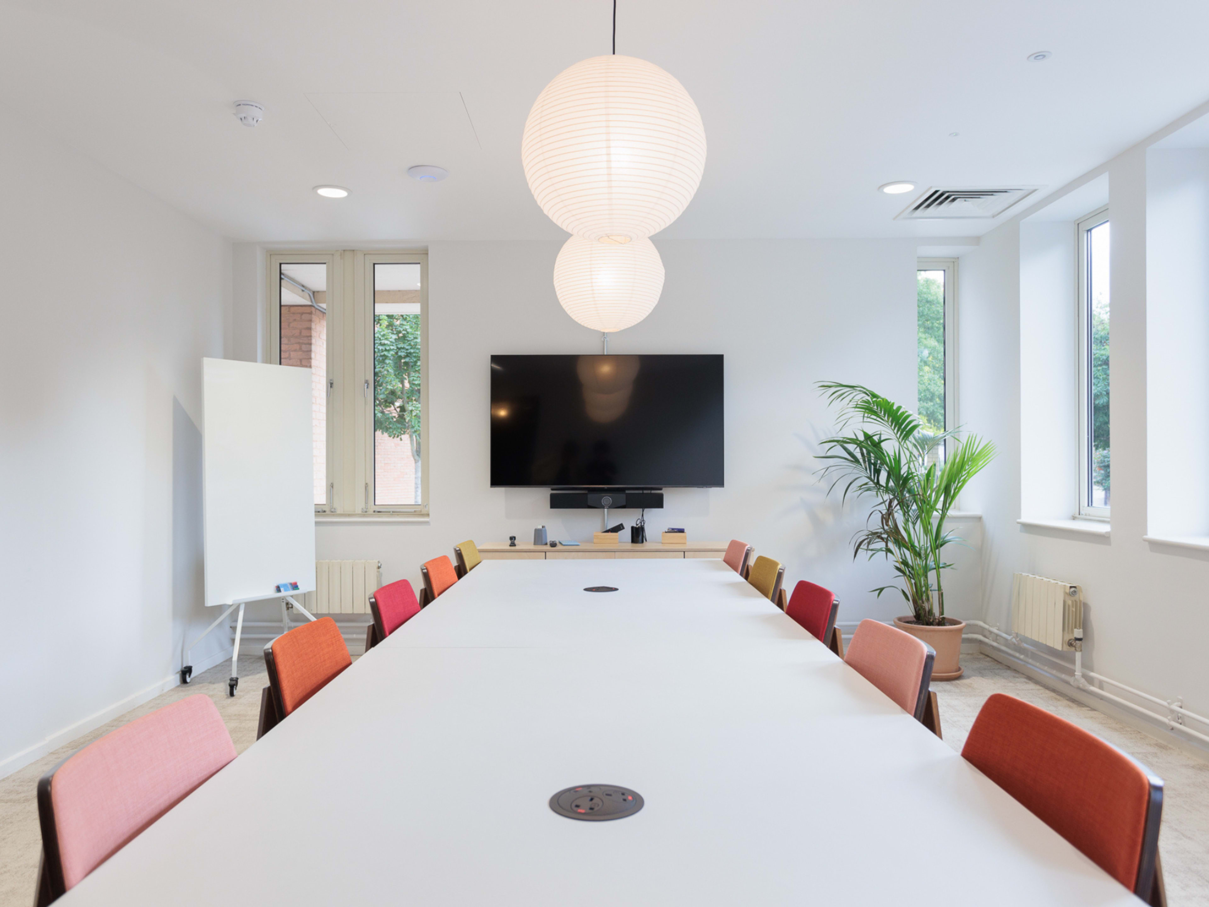 A modern conference room features a long table surrounded by colorful chairs, with a large television and a whiteboard on one wall.