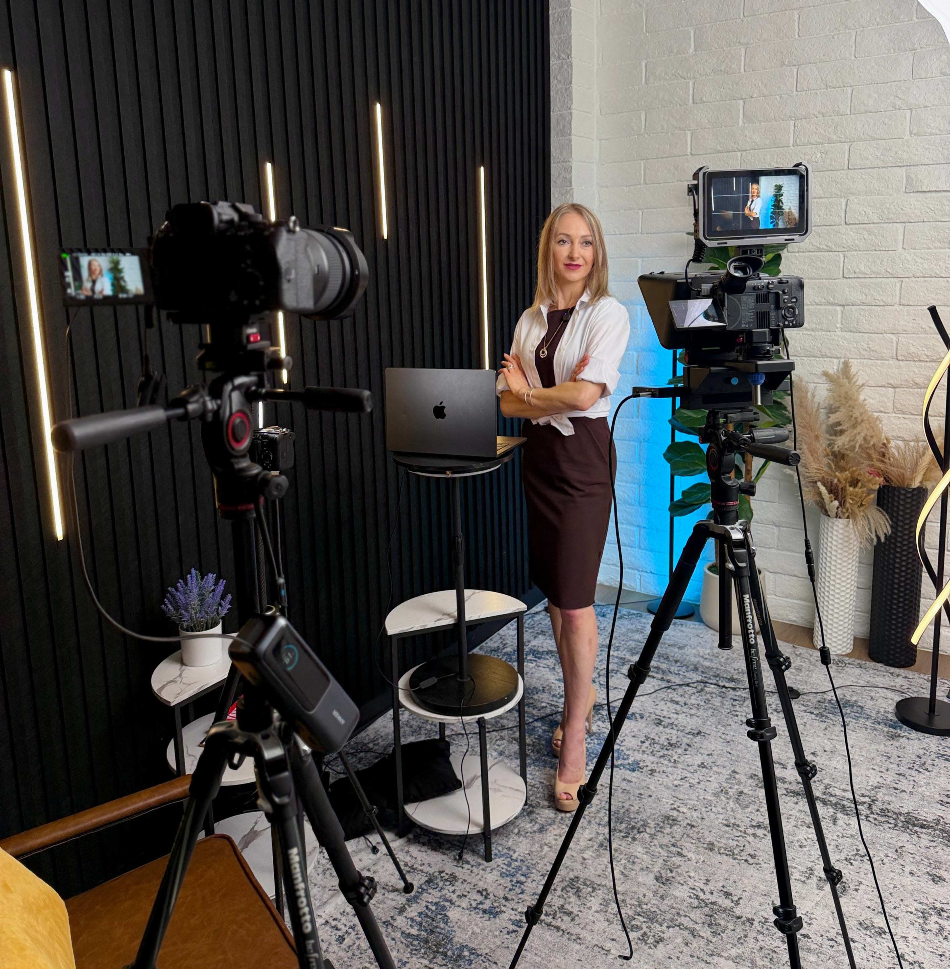A woman in a dark dress stands confidently beside a computer, with cameras and equipment set up for a video shoot in a well-lit studio.
