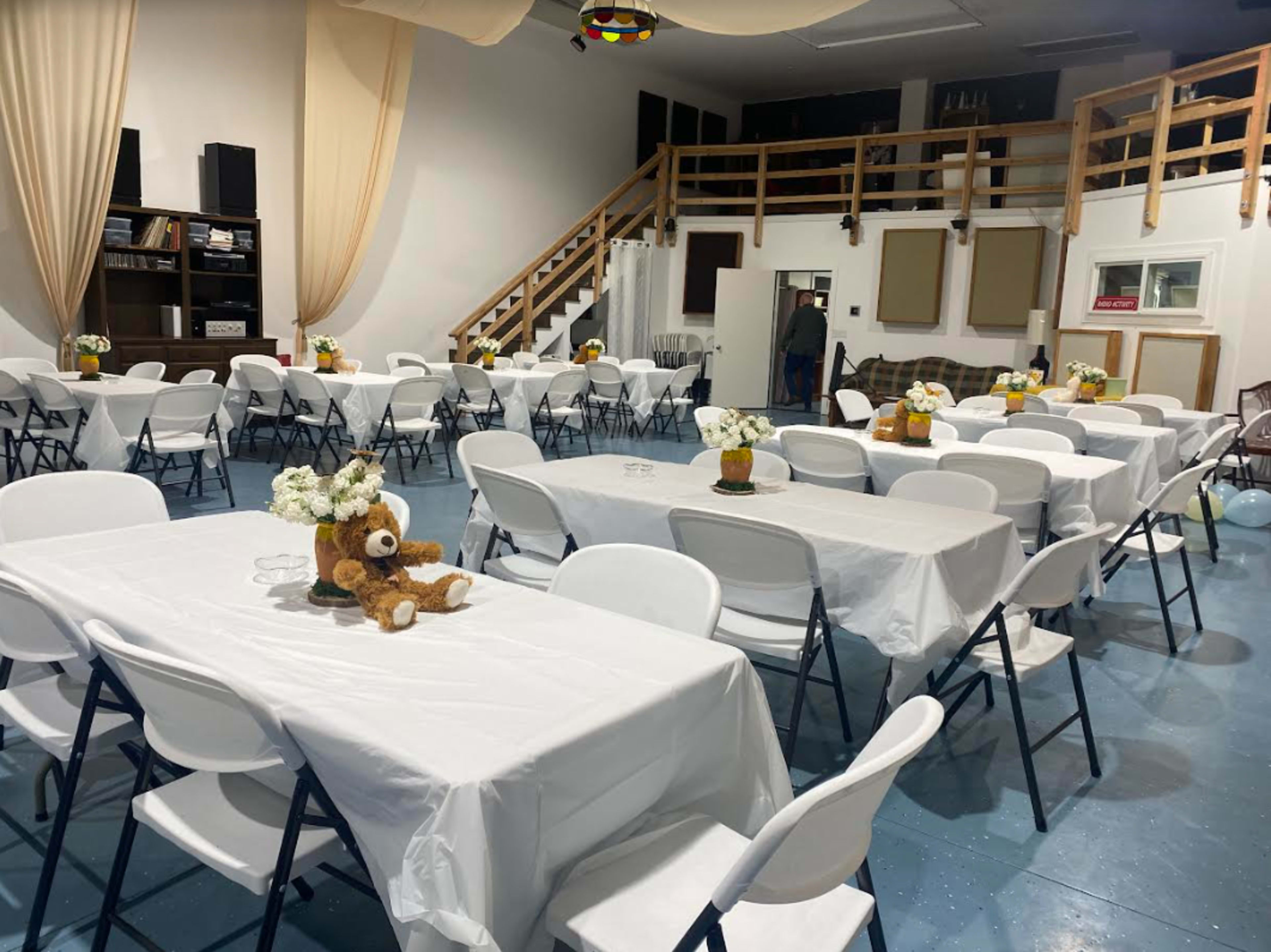 A large event space features multiple tables covered with white tablecloths, adorned with floral centerpieces, and a stuffed bear on one table.