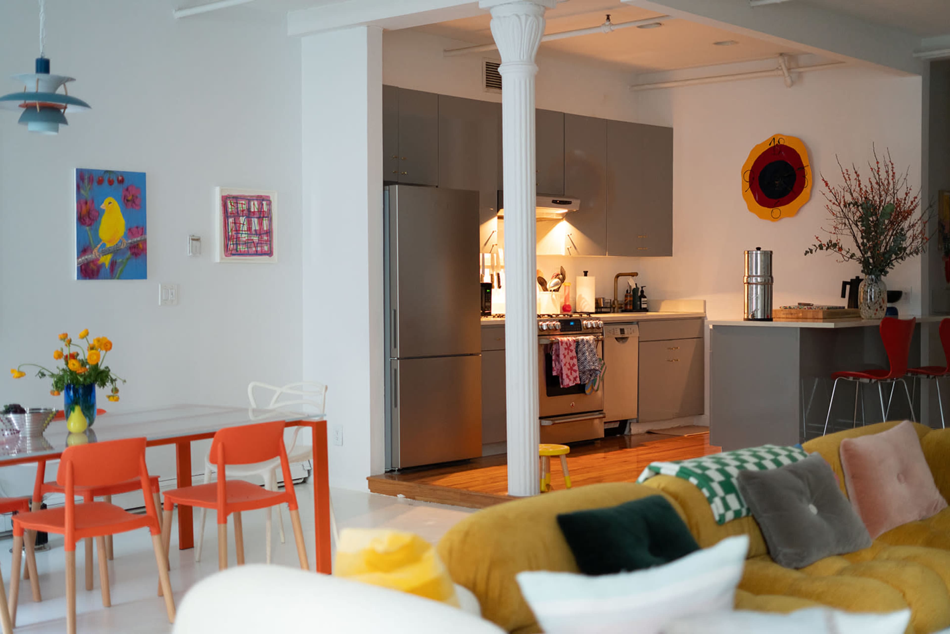 A modern kitchen with stainless steel appliances is visible through an open floor plan, featuring a dining area with colorful chairs and a brightly colored couch in the foreground.