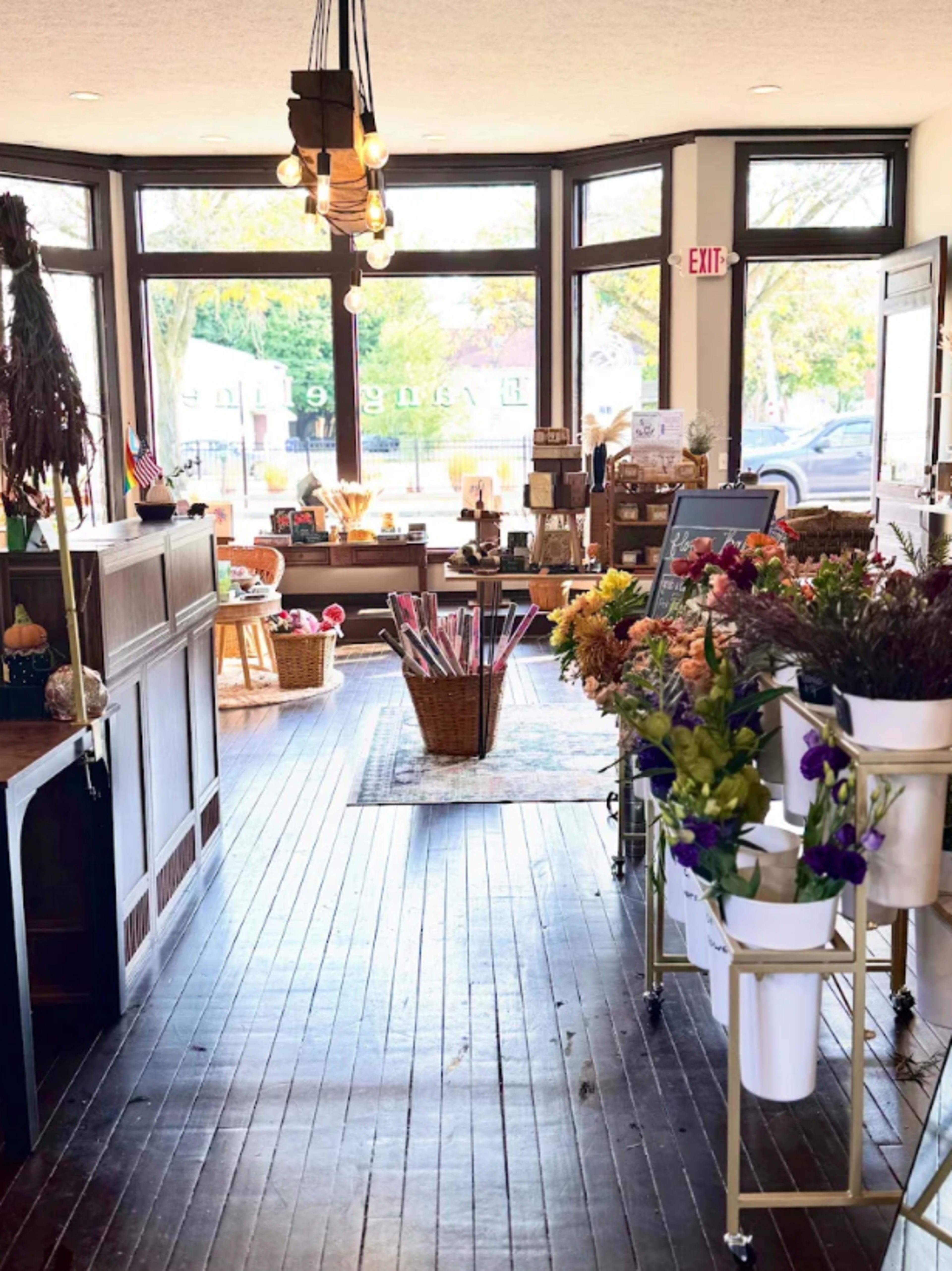 The image shows the interior of a charming shop featuring a wooden floor, large windows, and displays of flowers and various items for sale.