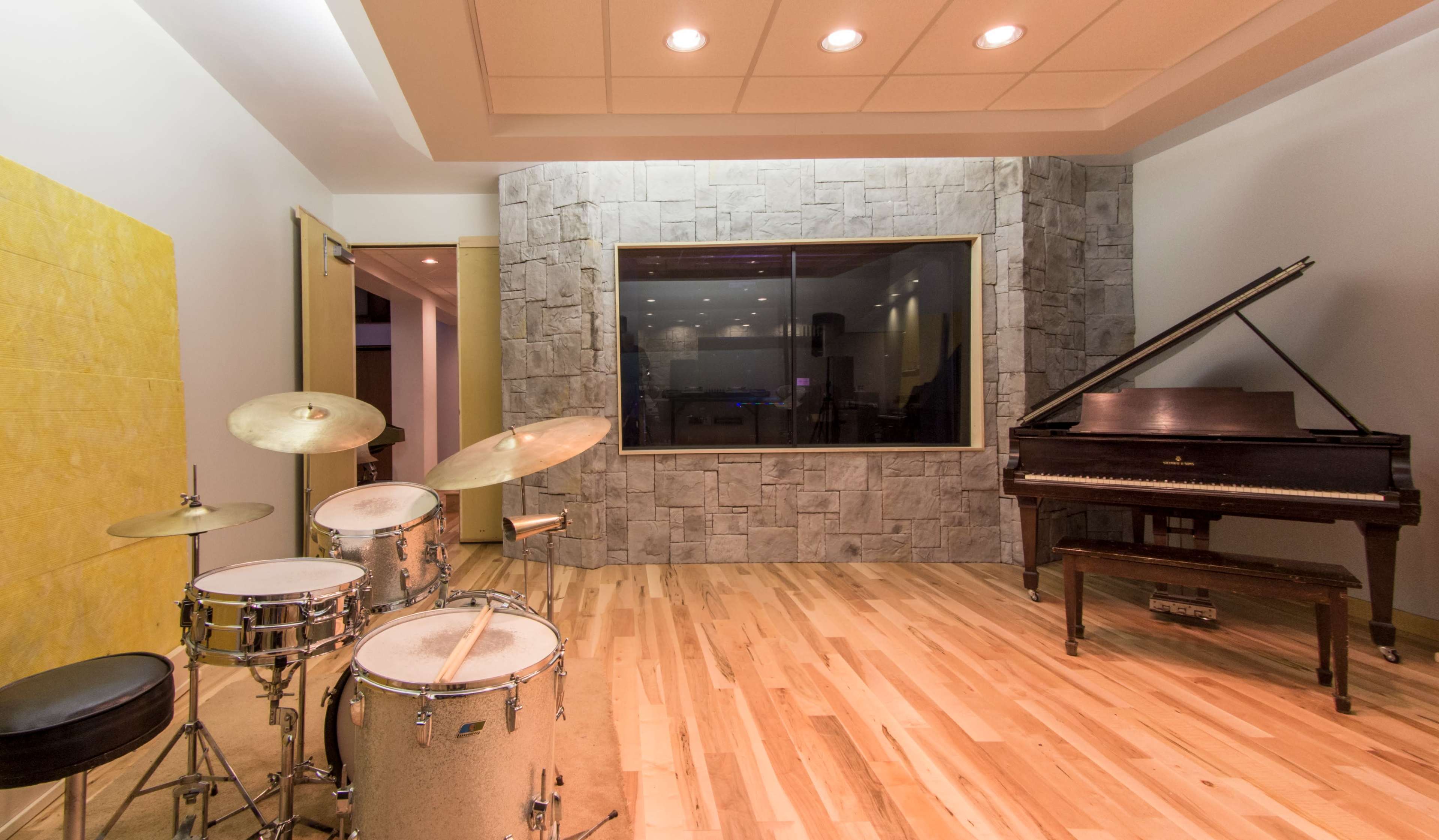 A music studio featuring a drum set and an upright piano, with large windows and stone accent walls.