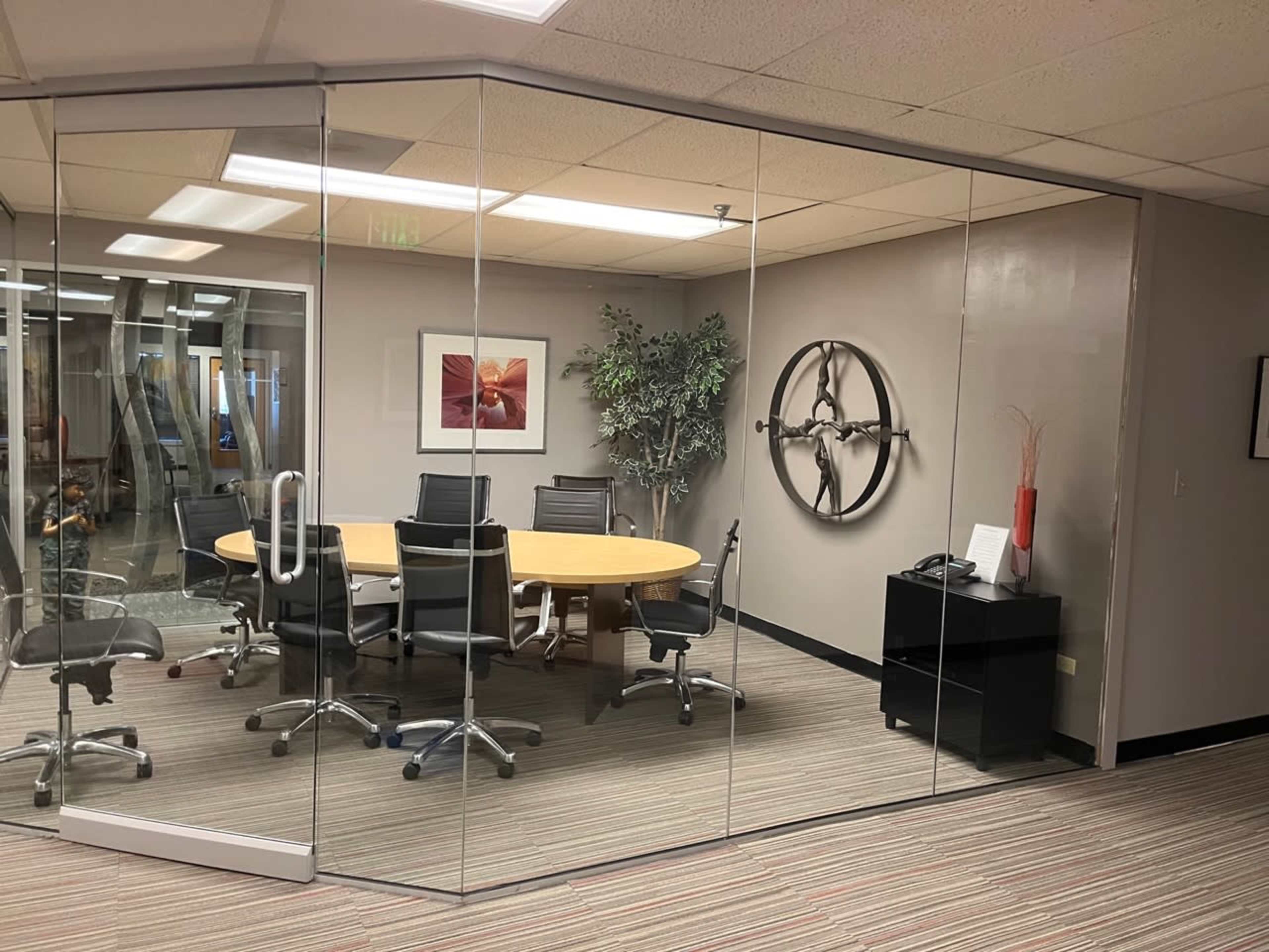 The image shows a glass-walled conference room with a circular table surrounded by black chairs, a plant in one corner, and a wall-mounted decoration.