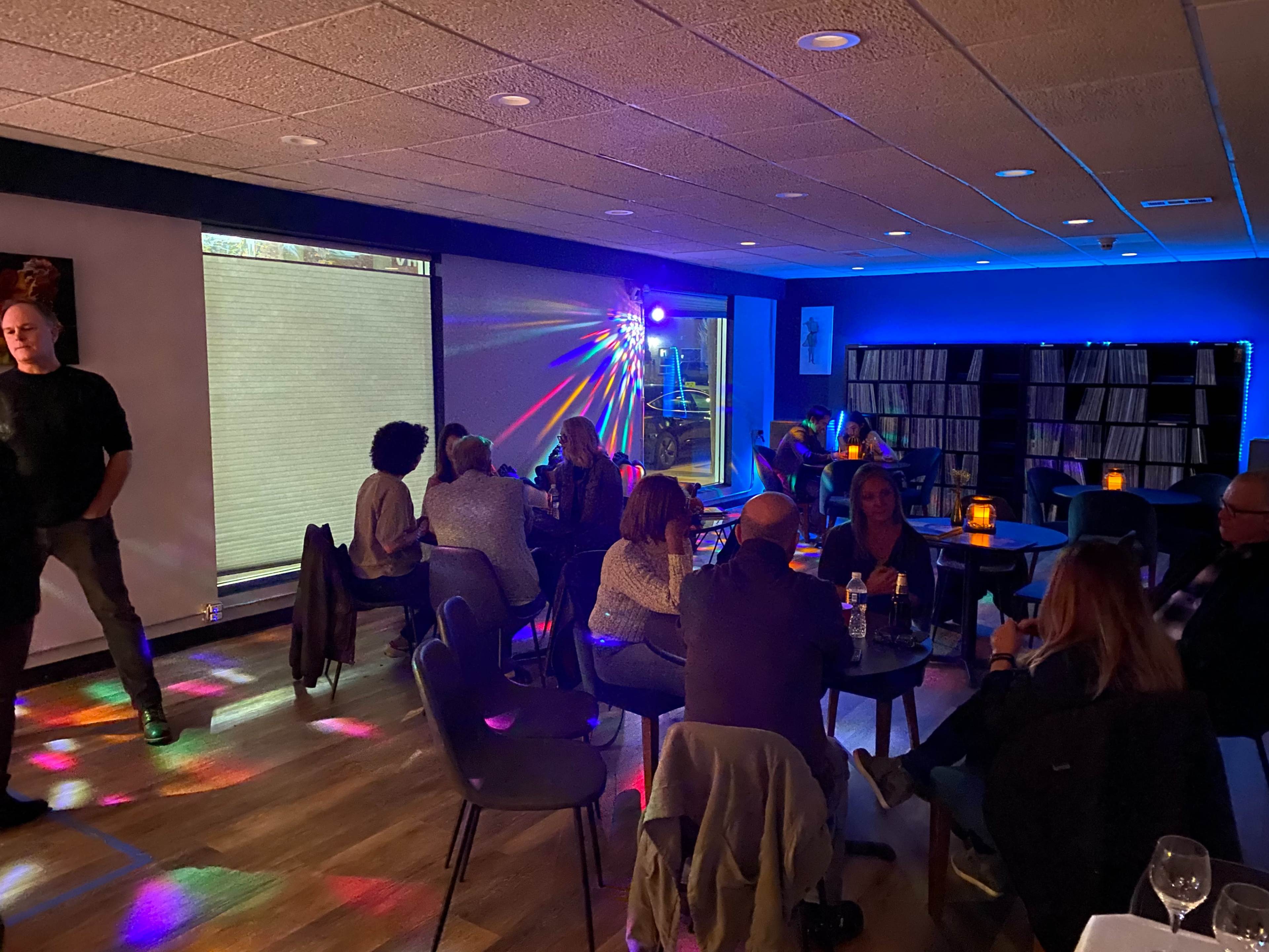 A dimly lit lounge features patrons seated at tables while colorful lights create patterns on the floor and walls.