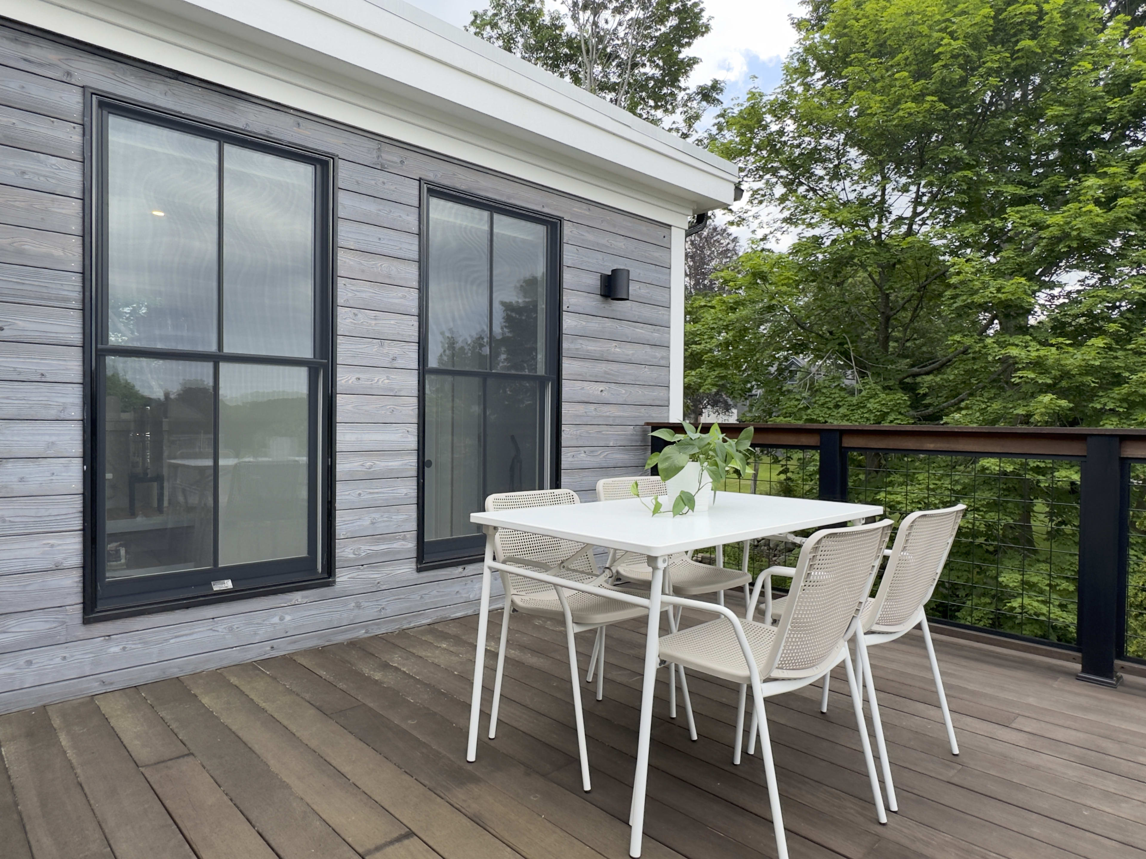 A modern outdoor dining area with a white table and six chairs on a wooden deck, adjacent to a house with grey clapboard siding and large windows.