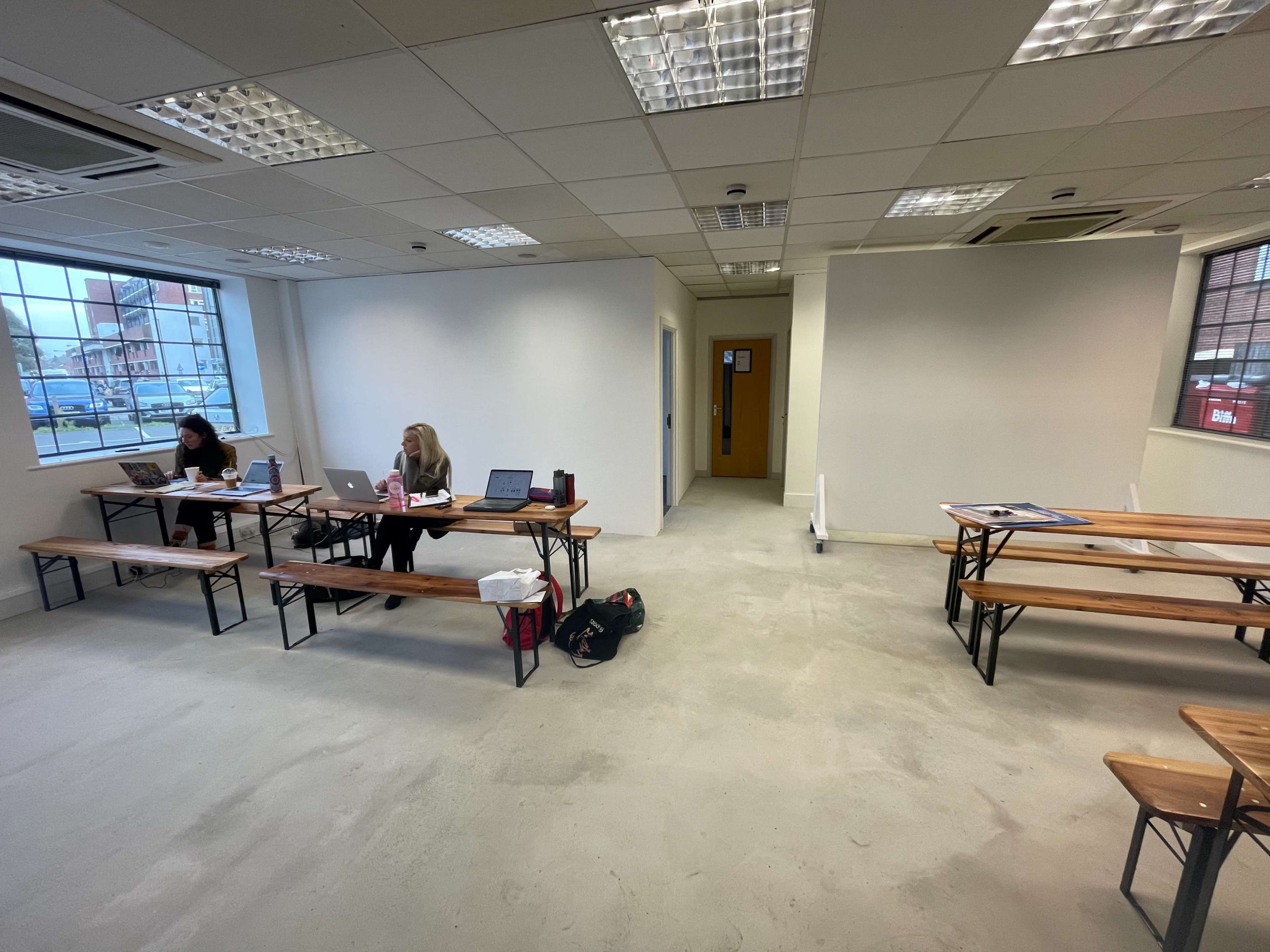 Two women sit at wooden tables with laptops in a sparsely furnished classroom featuring concrete floors and large windows.