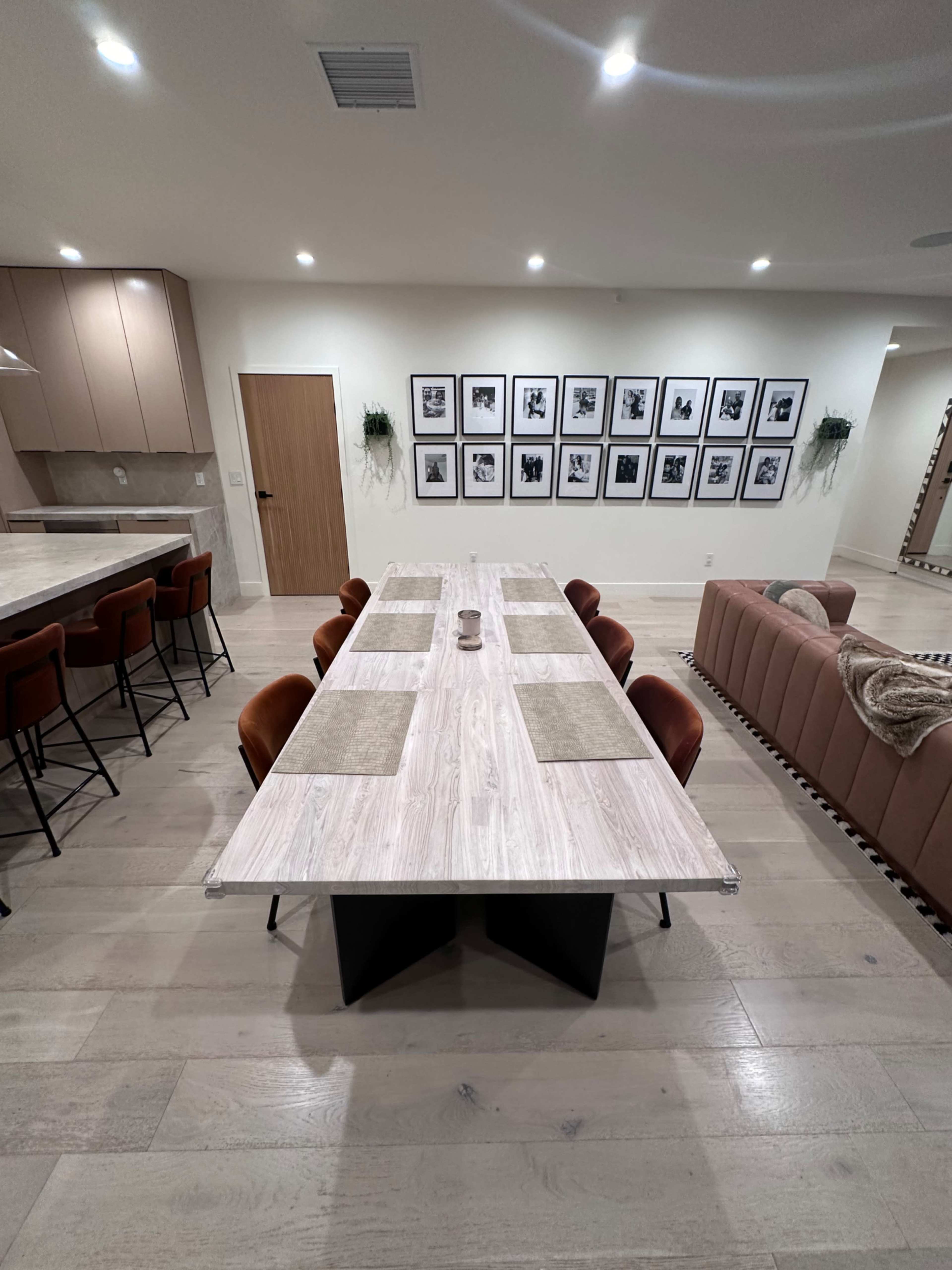 The image shows a modern dining area featuring a long table with rectangular placemats, surrounded by upholstered chairs, and a gallery wall of framed photos in the background.