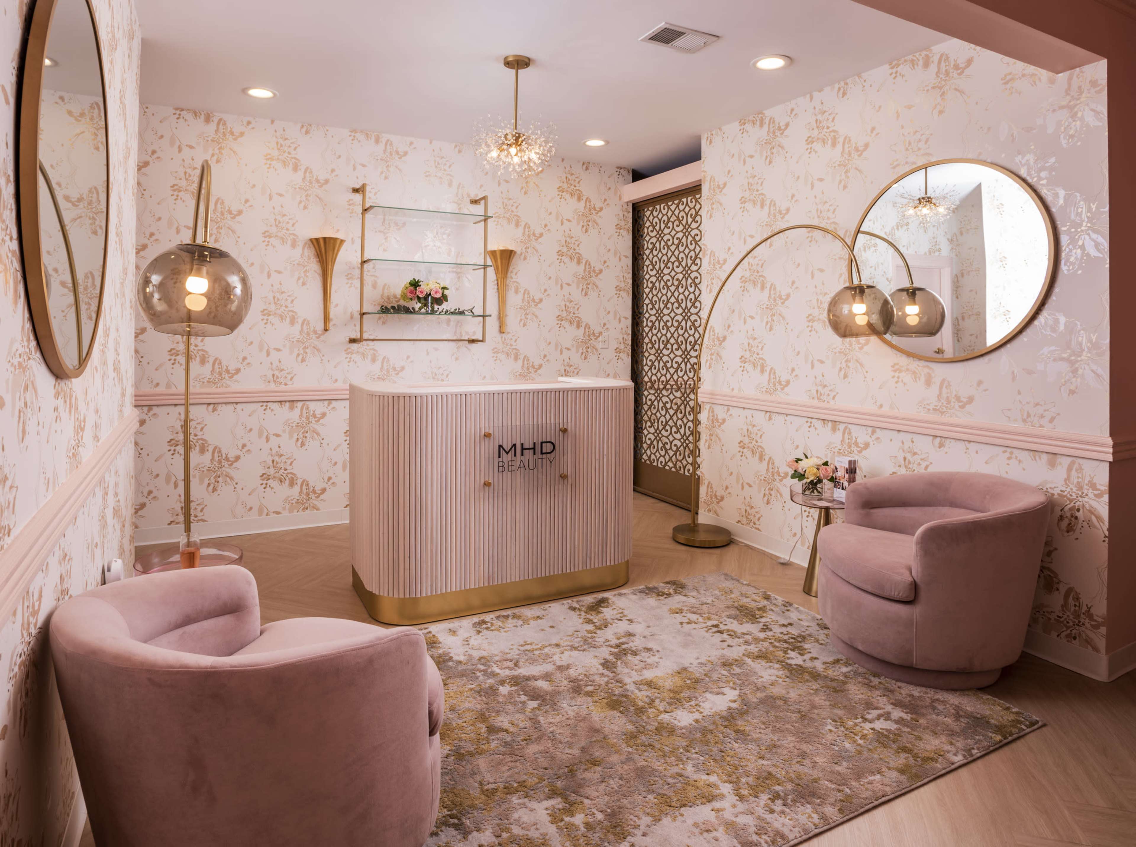 A modern beauty salon reception area featuring a curved front desk, two plush armchairs, and decorative wall mirrors.