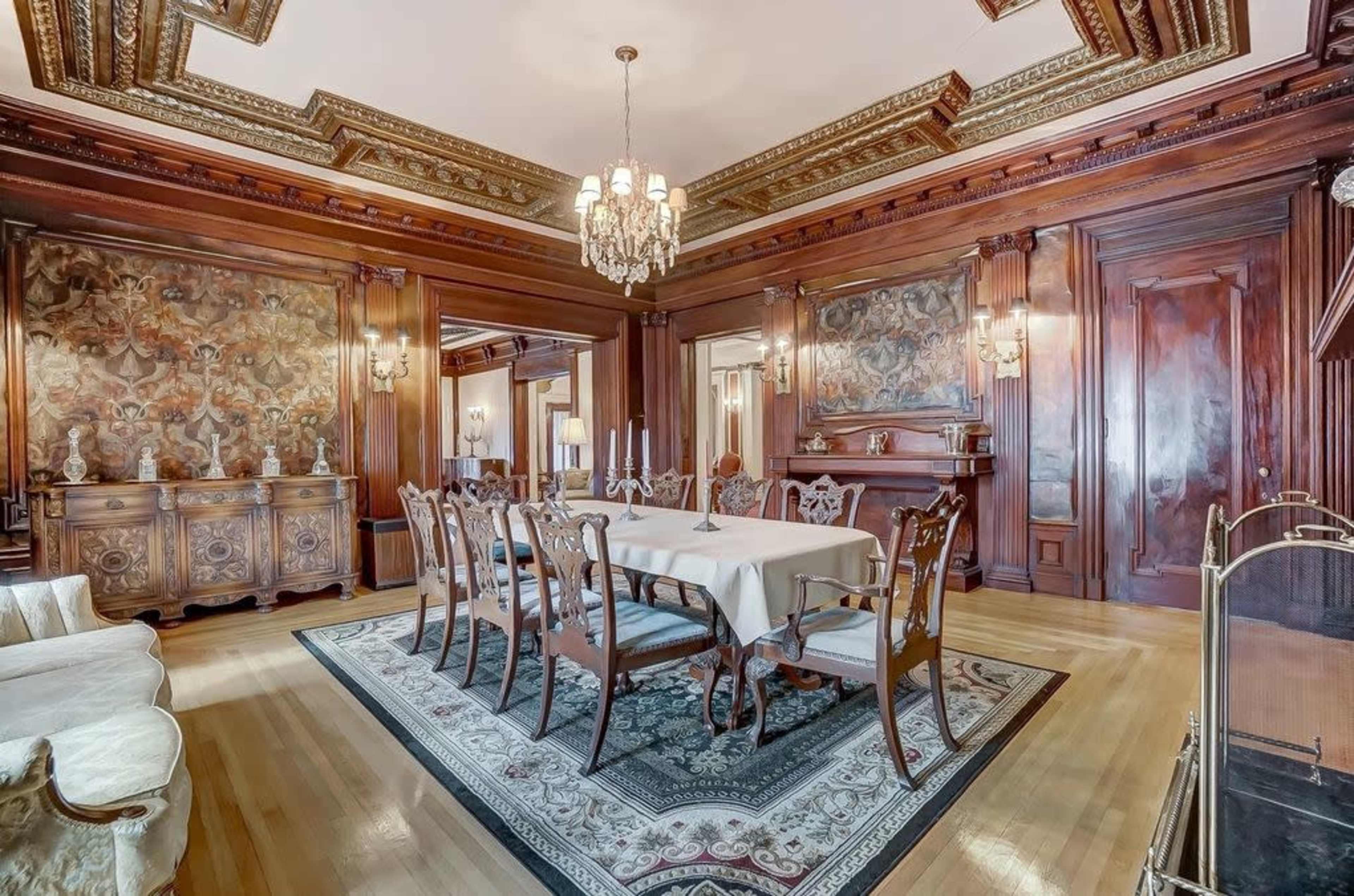 An elegant dining room with a large table surrounded by ornate chairs, featuring detailed wood paneling and a chandelier overhead.