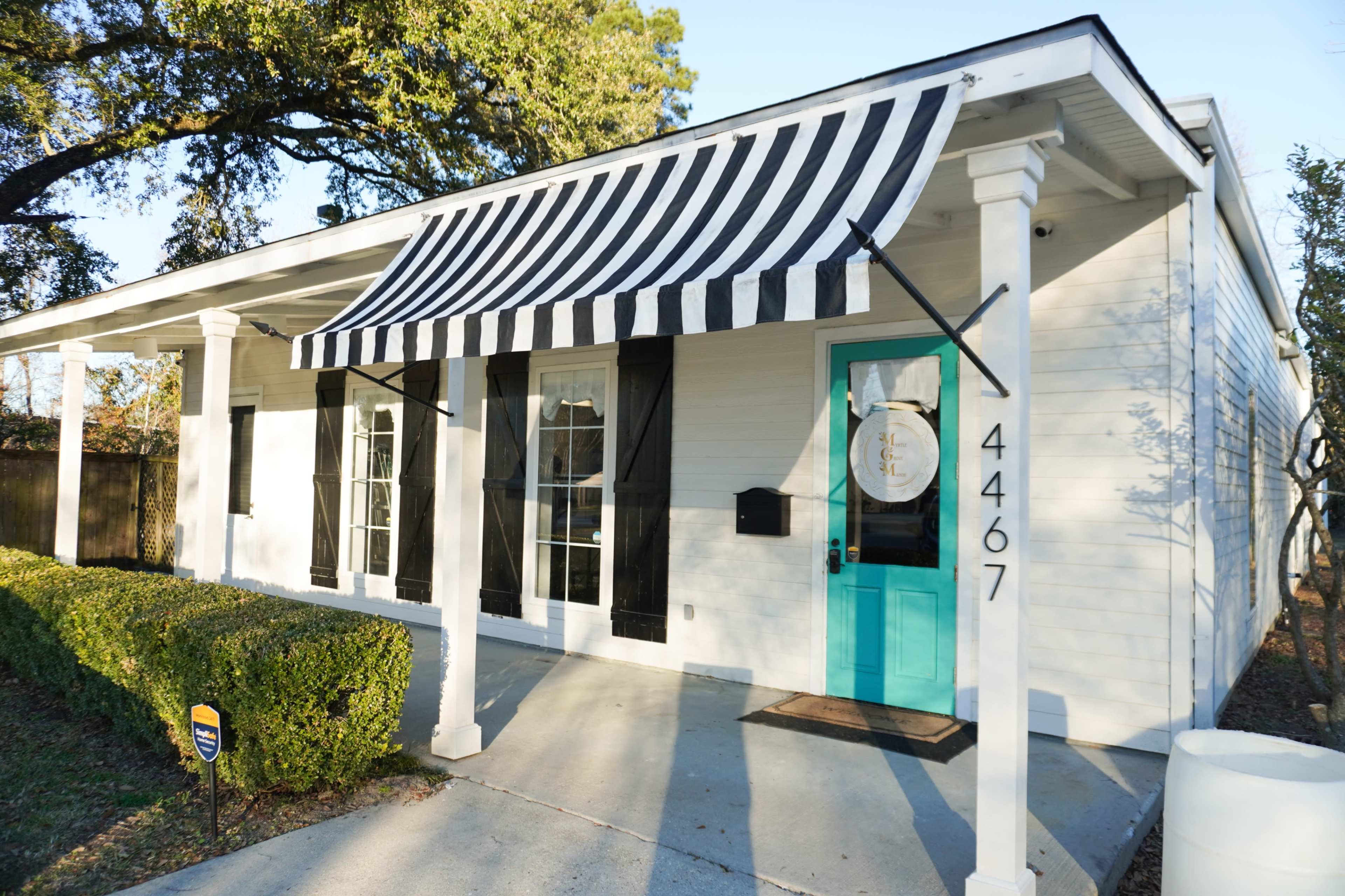 The image shows a white house with a black and white striped awning, a turquoise front door, and neatly trimmed shrubs in front.