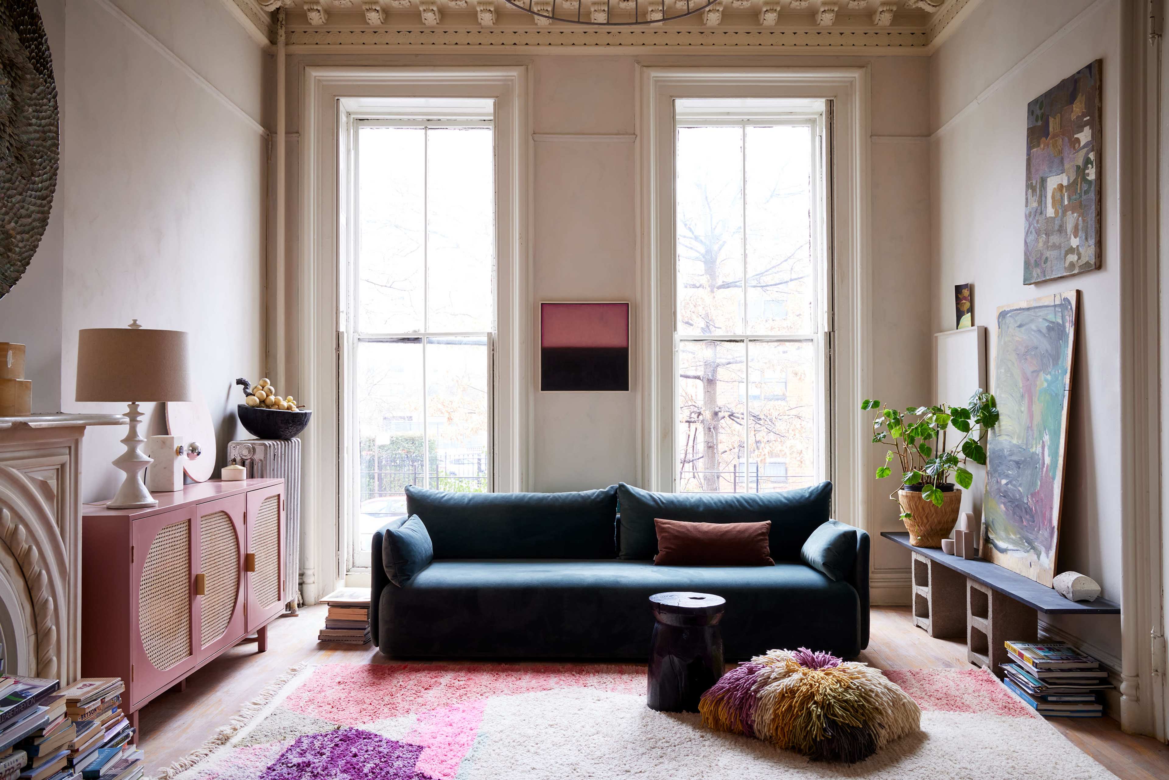 The living room features a dark green sofa, a pink sideboard, large windows with natural light, and decor that includes plants and artwork on the walls.