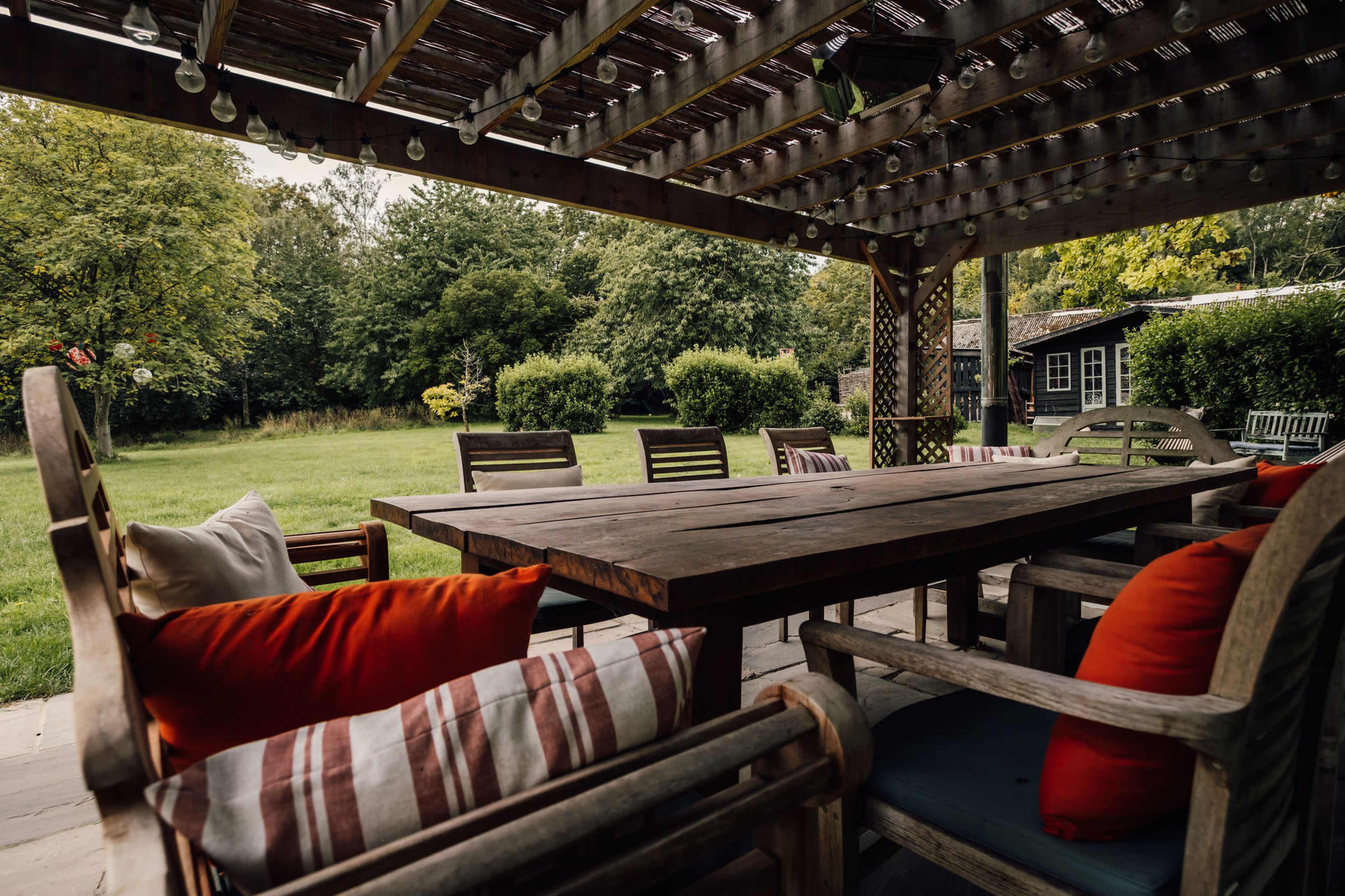 A wooden patio table with colorful cushions is set beneath a pergola, overlooking a grassy yard surrounded by trees and shrubs.