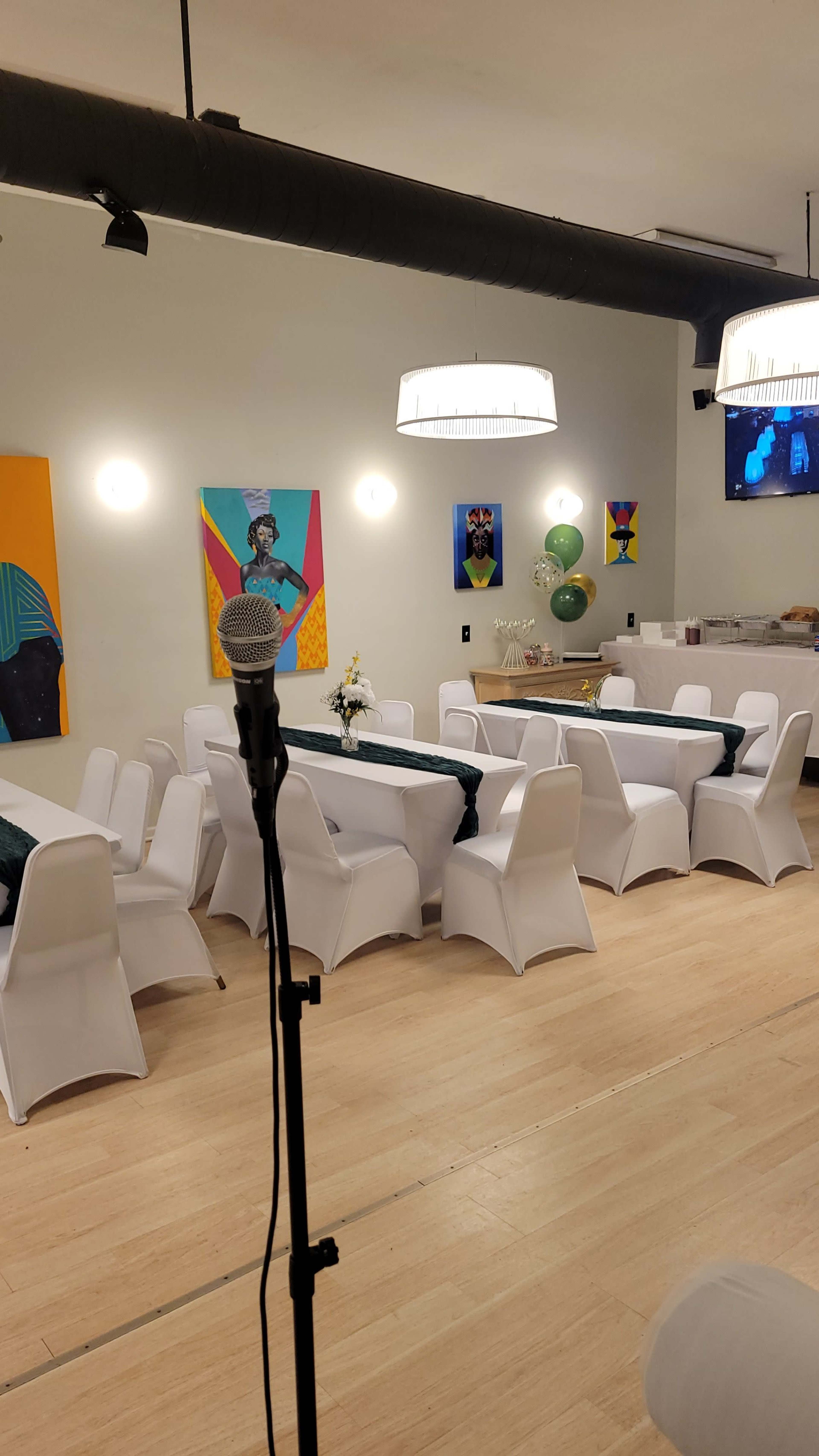 The image shows a spacious event room with white tables and chairs arranged for a gathering, featuring colorful artwork on the walls.