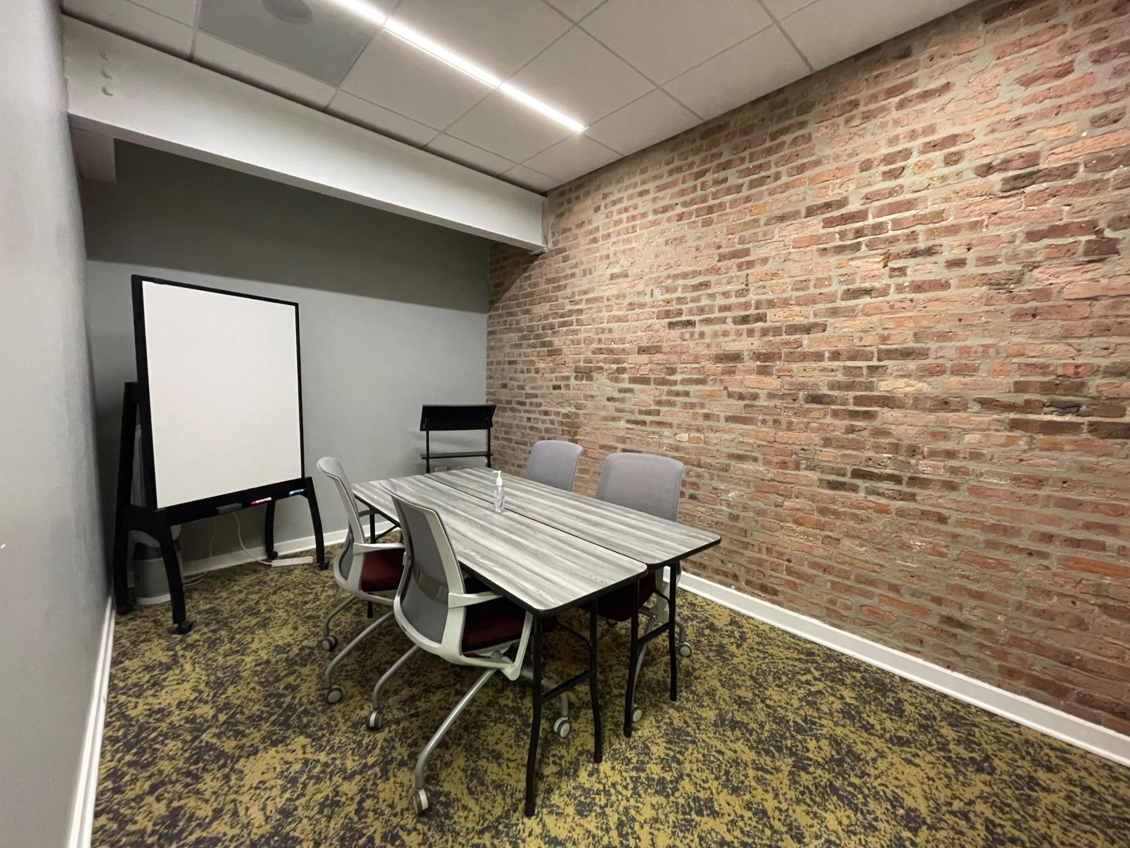 The image shows a small meeting room with a rectangular table, four chairs, a whiteboard, and walls made of exposed brick.