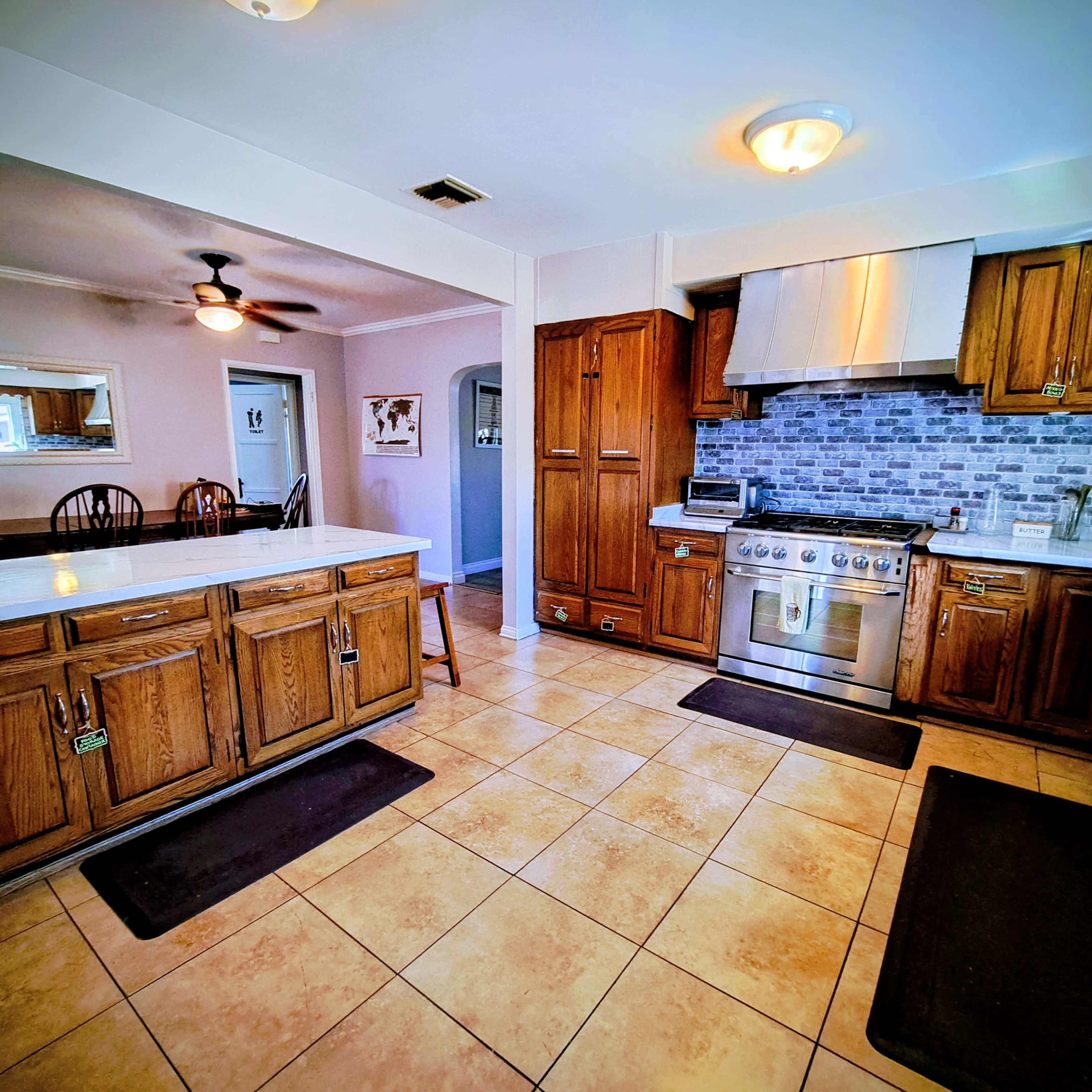 The image shows a kitchen with wooden cabinetry, a stainless steel stove, and tiled flooring, featuring a dining area visible in the background.