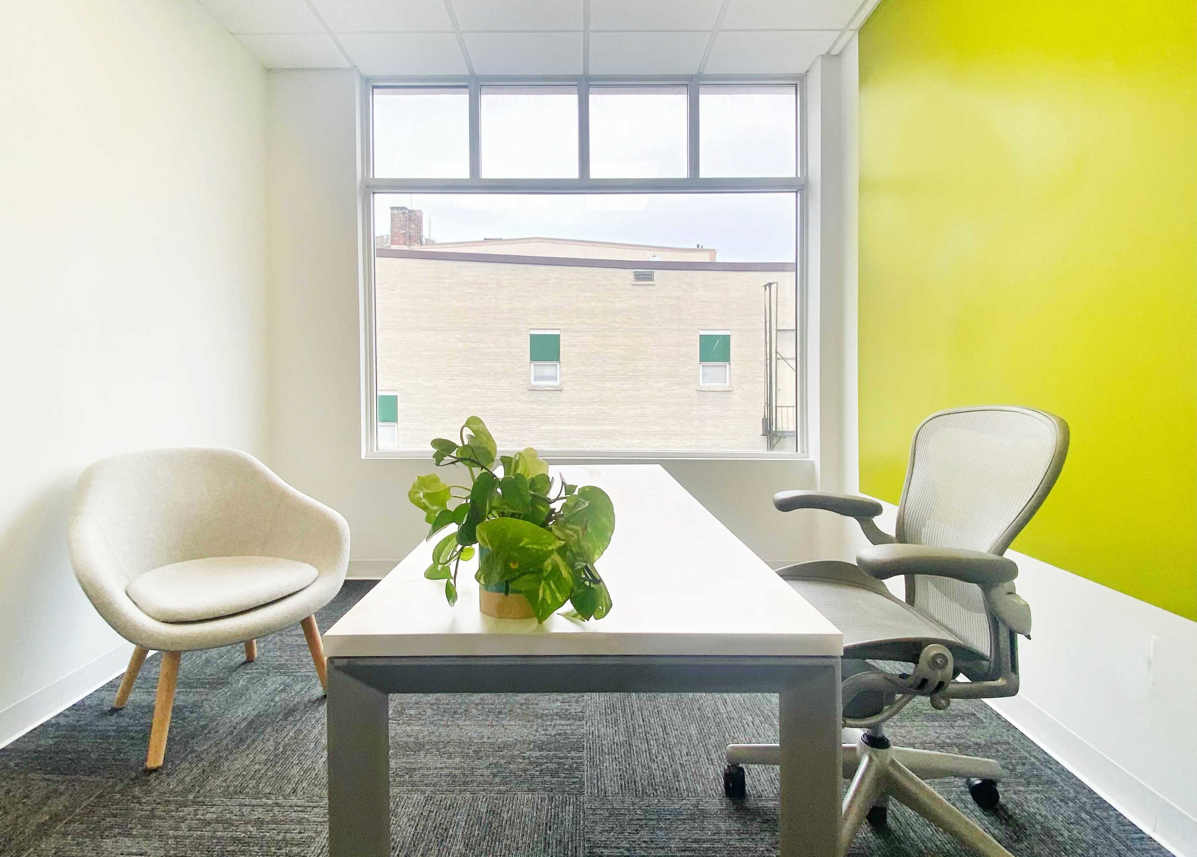 A modern office space featuring a table with a potted plant, a chair, and a window overlooking a building outside.