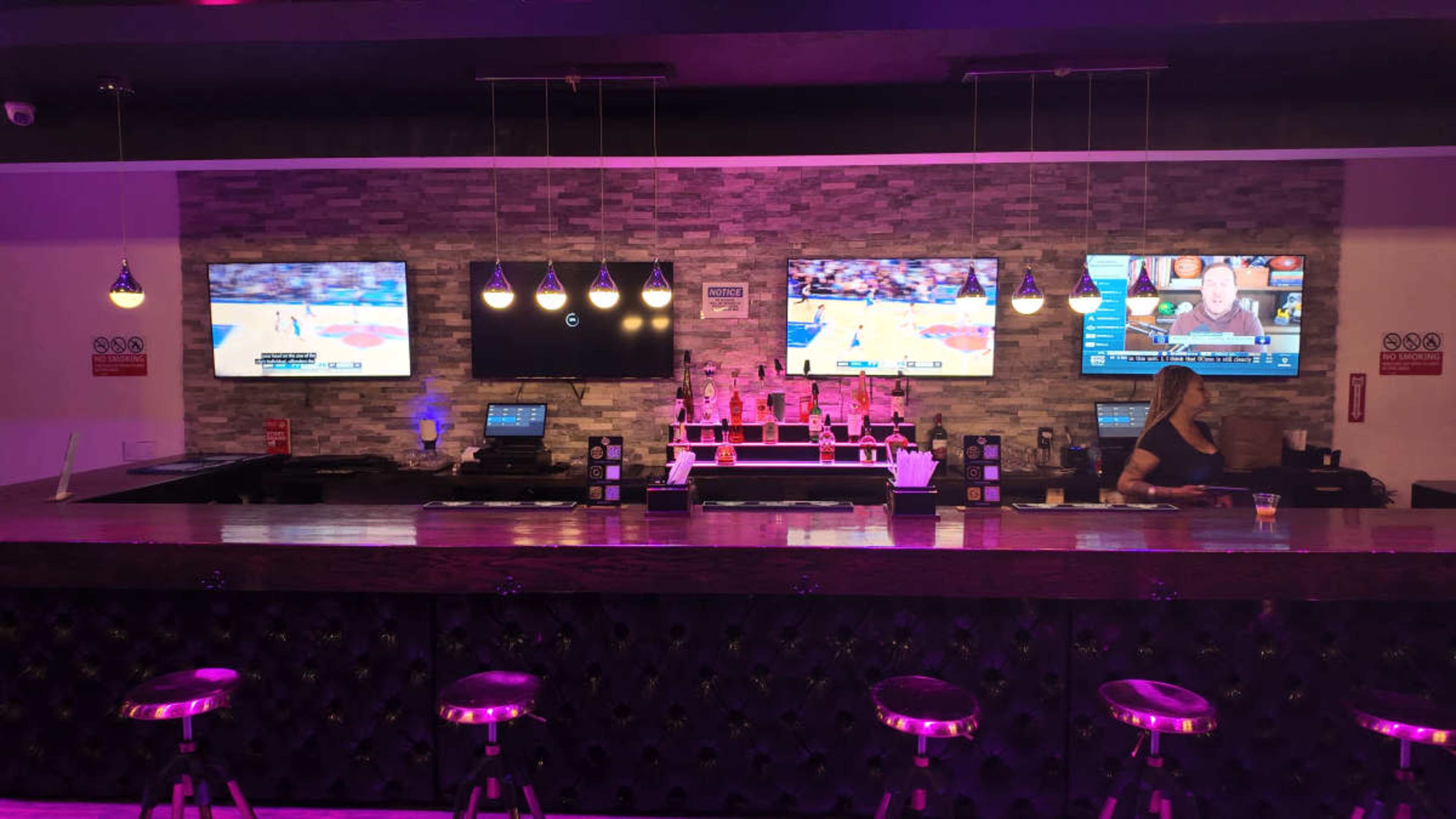 The image shows a modern bar with a wooden counter, multiple televisions displaying sports, and a bartender preparing drinks in a well-lit environment.