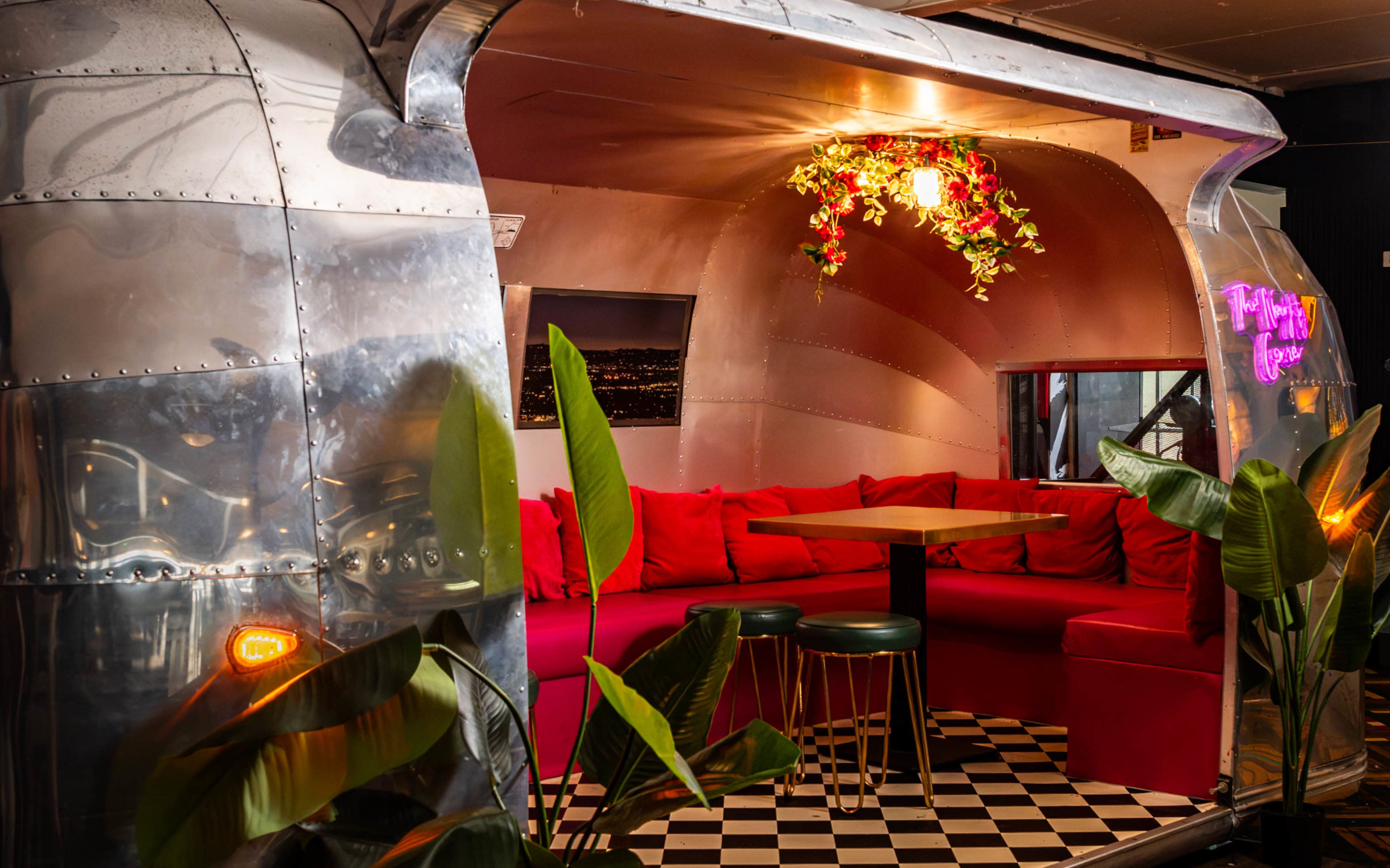 The interior features a rounded, metallic booth adorned with red seating and a small table, surrounded by green plants and a checkered floor.