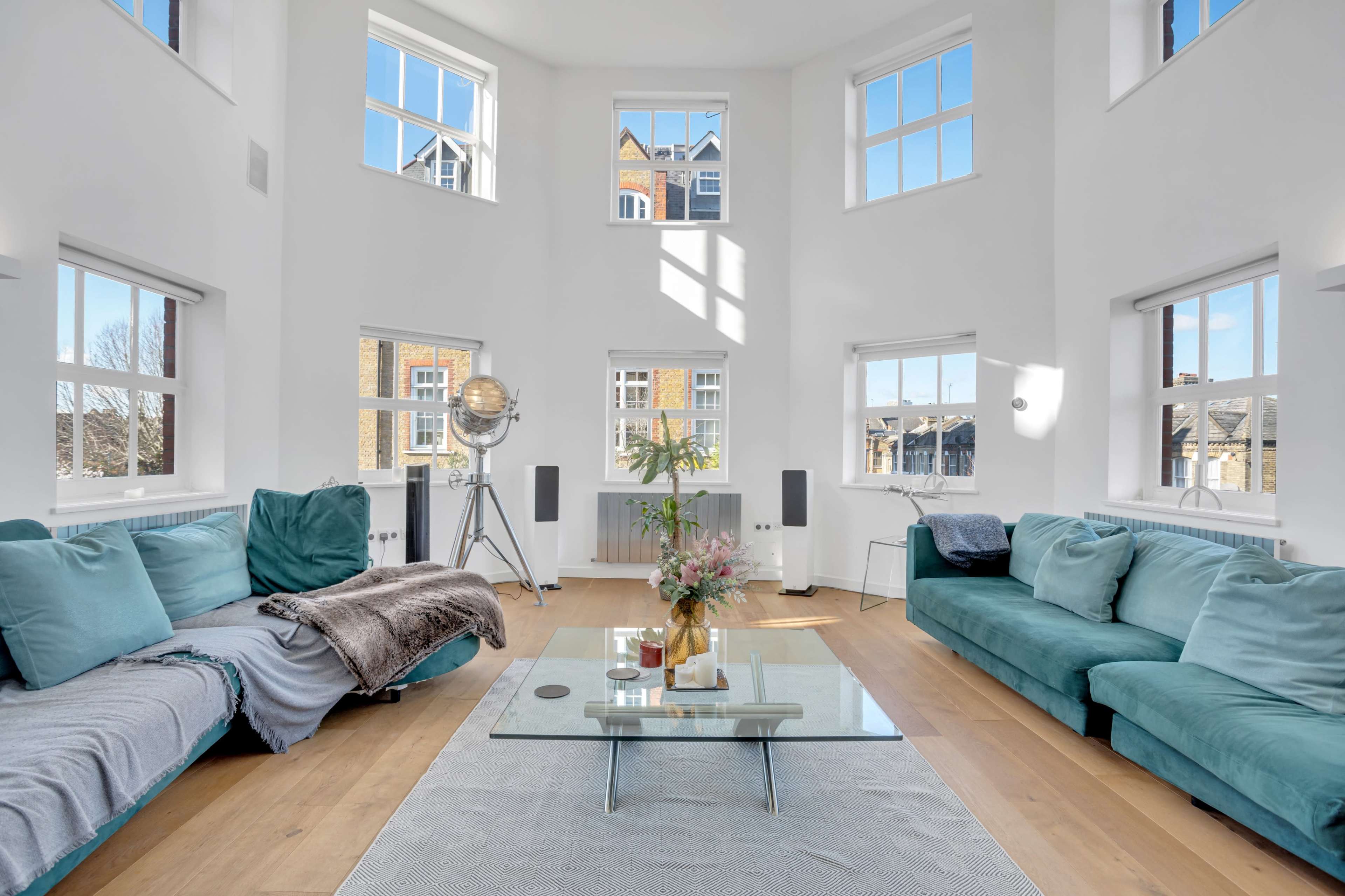 A spacious living room features turquoise sofas, a glass coffee table, and large windows that allow natural light to fill the space.