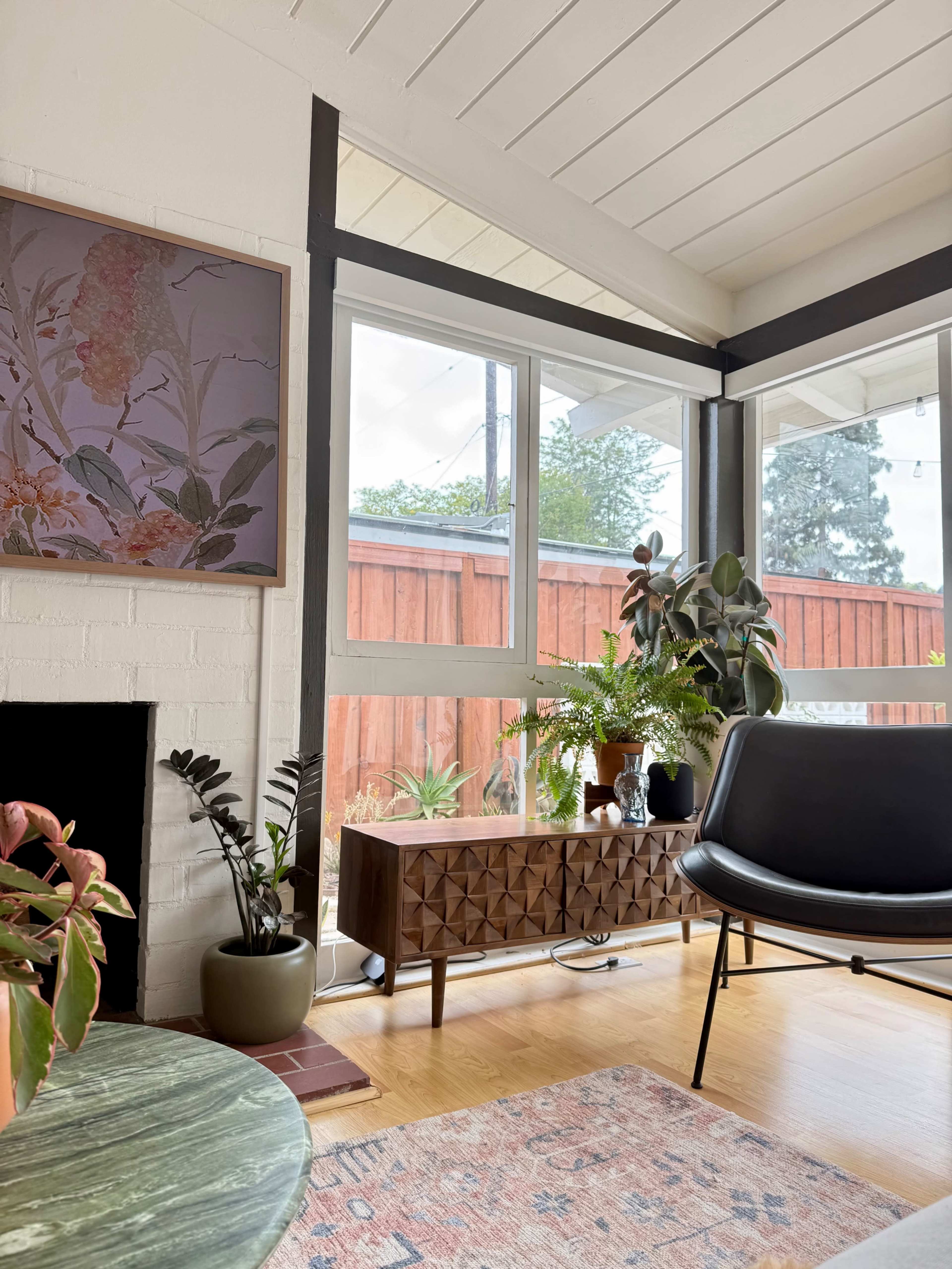 The image shows a cozy interior with a modern black chair, a wooden sideboard, potted plants, and large windows providing natural light.