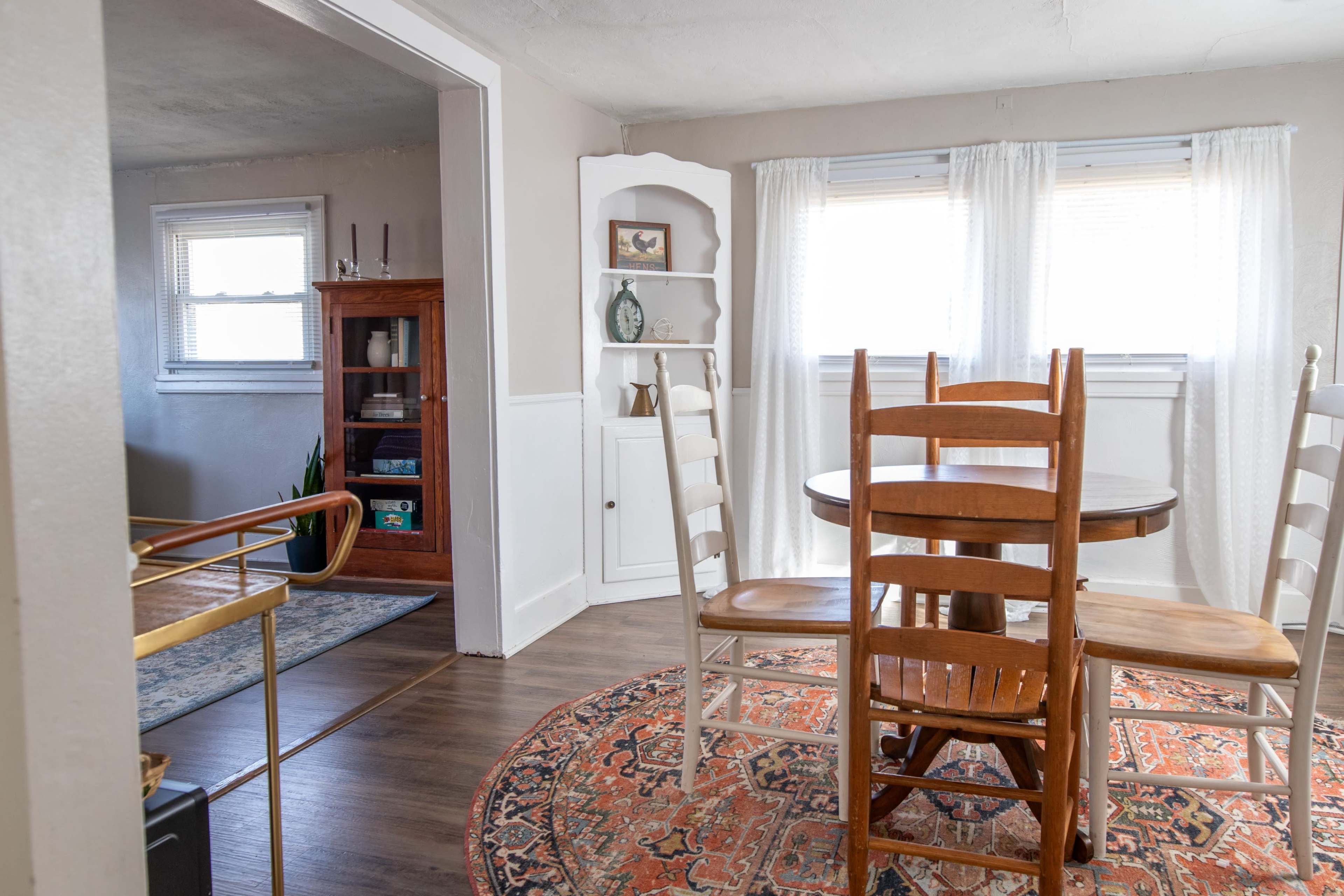 A wooden dining table with chairs is placed on a patterned rug in a well-lit room, with a doorway leading to another area featuring a wooden cabinet and shelves.