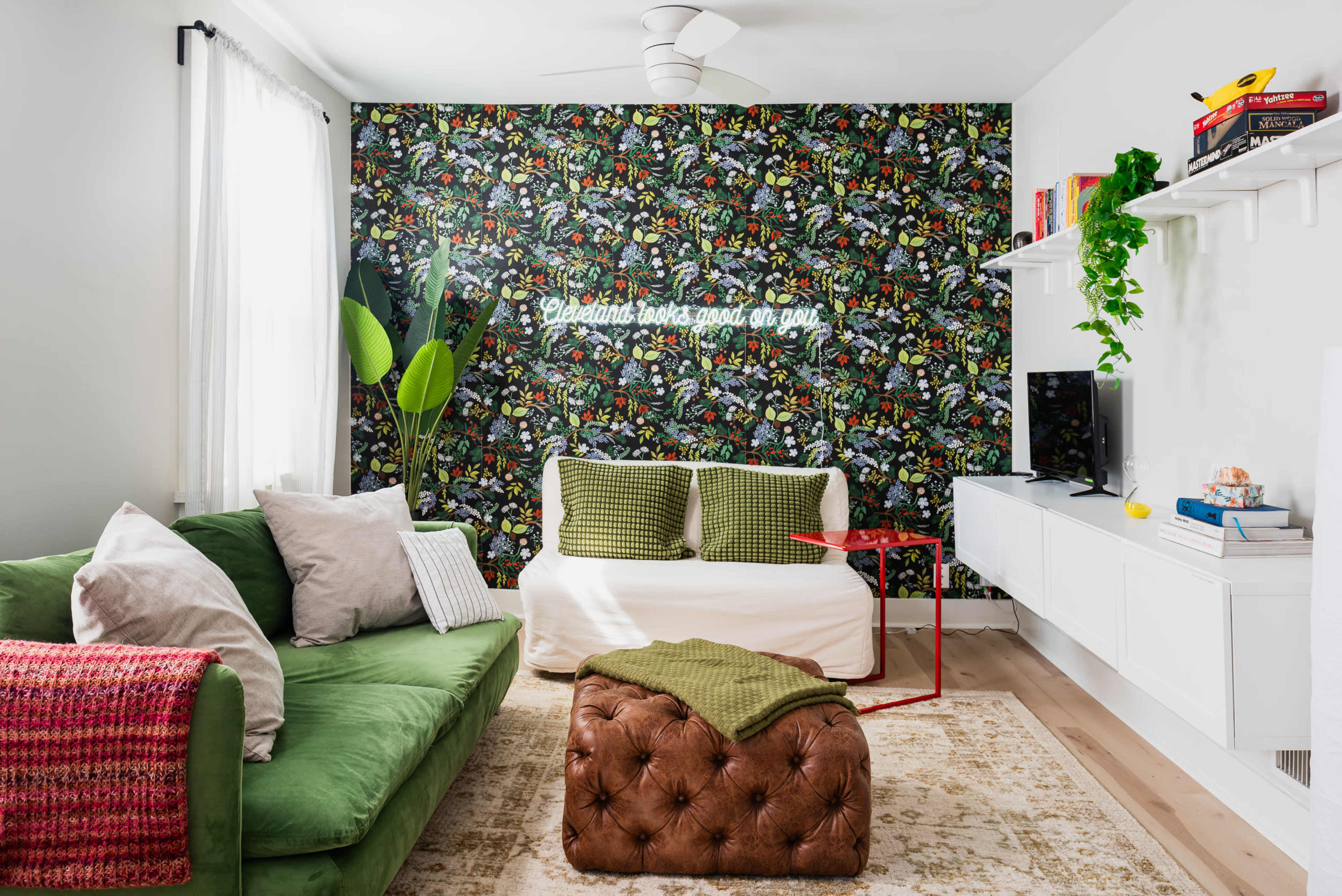 A modern living room features a green sofa, a white couch, a brown tufted ottoman, and a colorful accent wall with a neon sign, complemented by a television and decorative shelves.