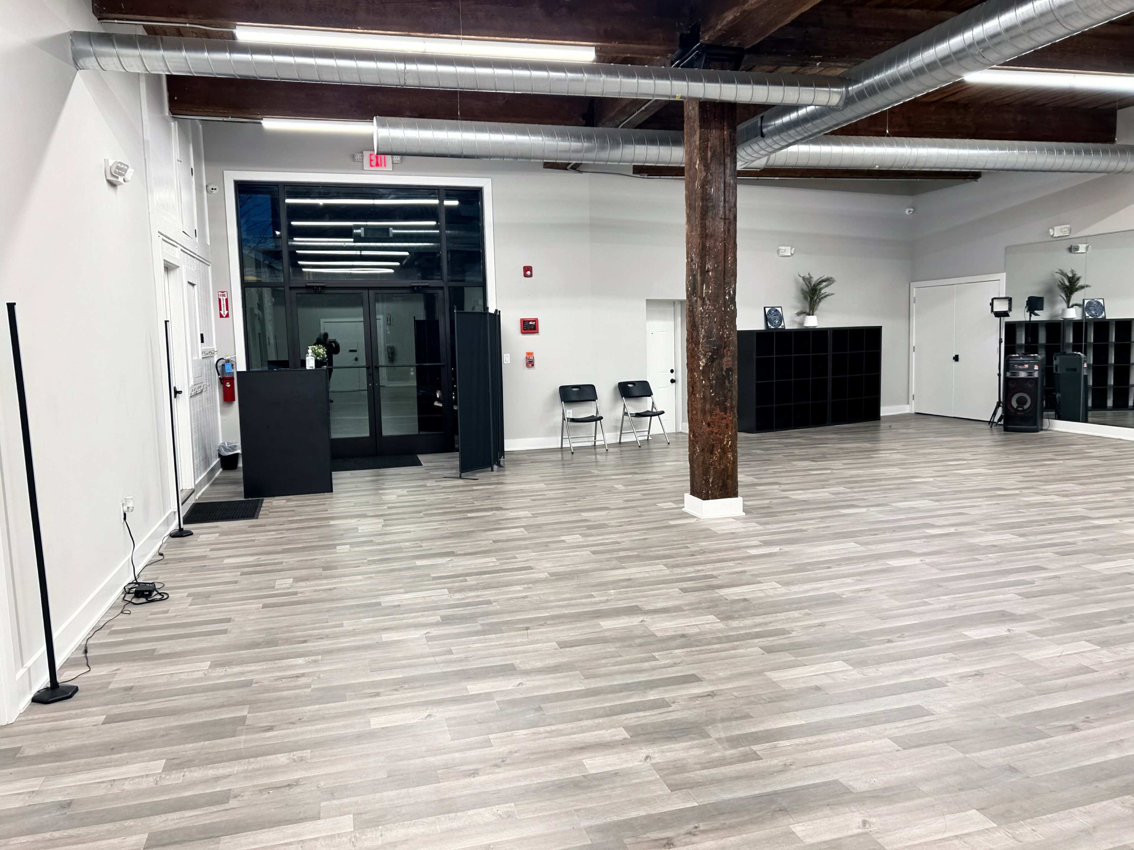 The image shows a spacious interior room with wooden flooring, large windows, and a few chairs positioned near a black partition.