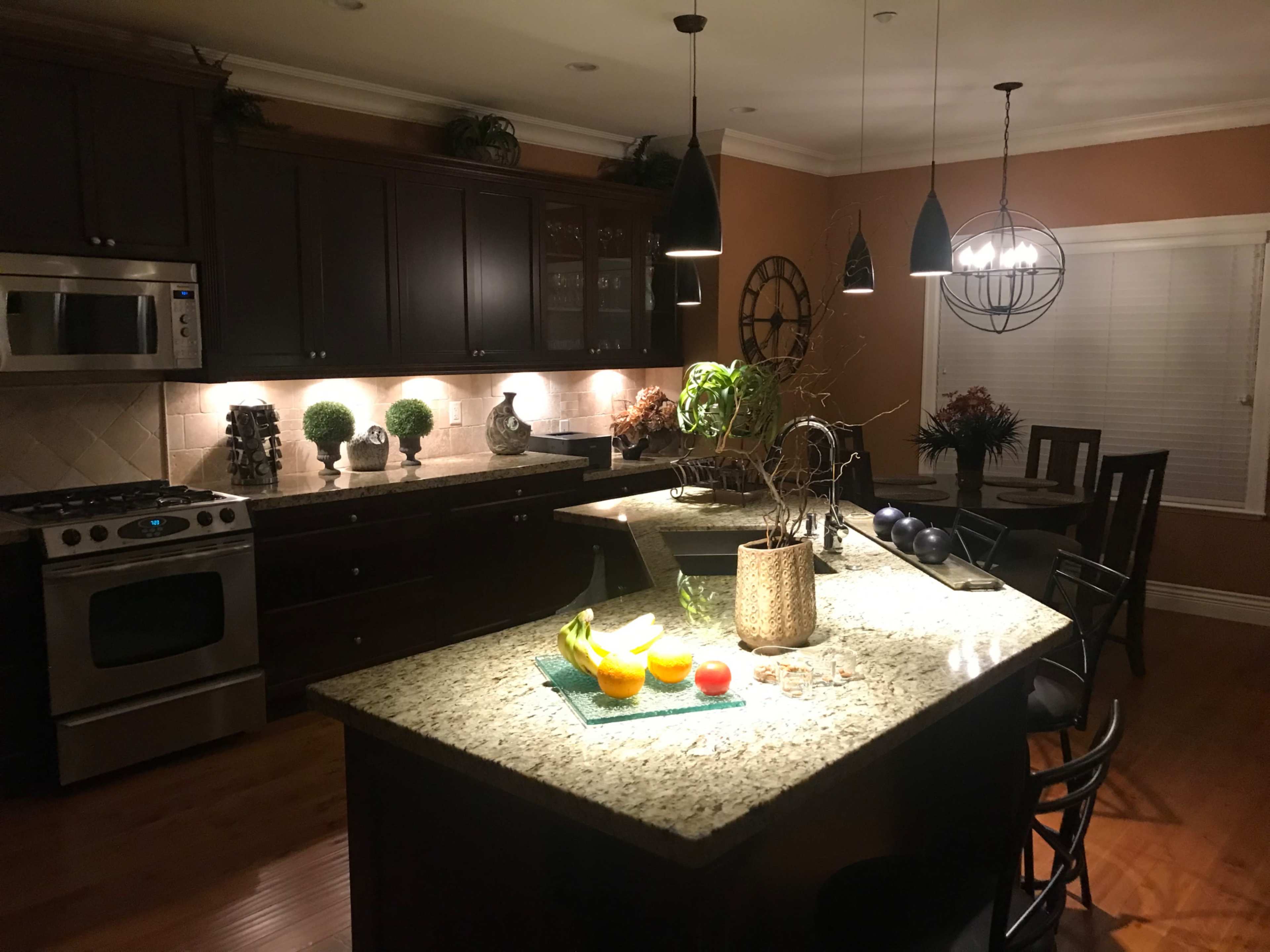 A kitchen with dark cabinetry, a granite island, and ambient lighting, featuring a bowl of fruits on the countertop.