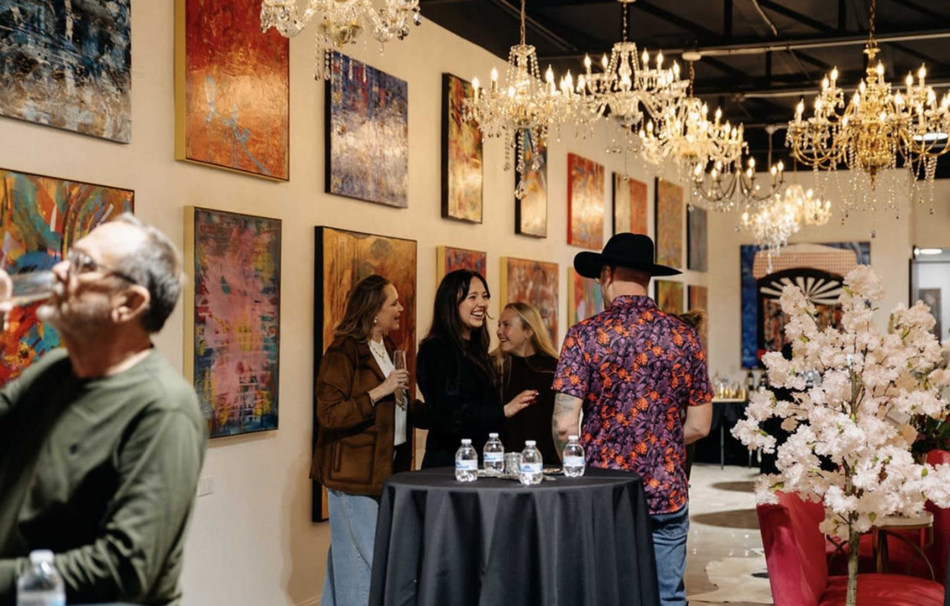 A group of people socialize in a well-lit art gallery featuring colorful paintings and chandeliers.