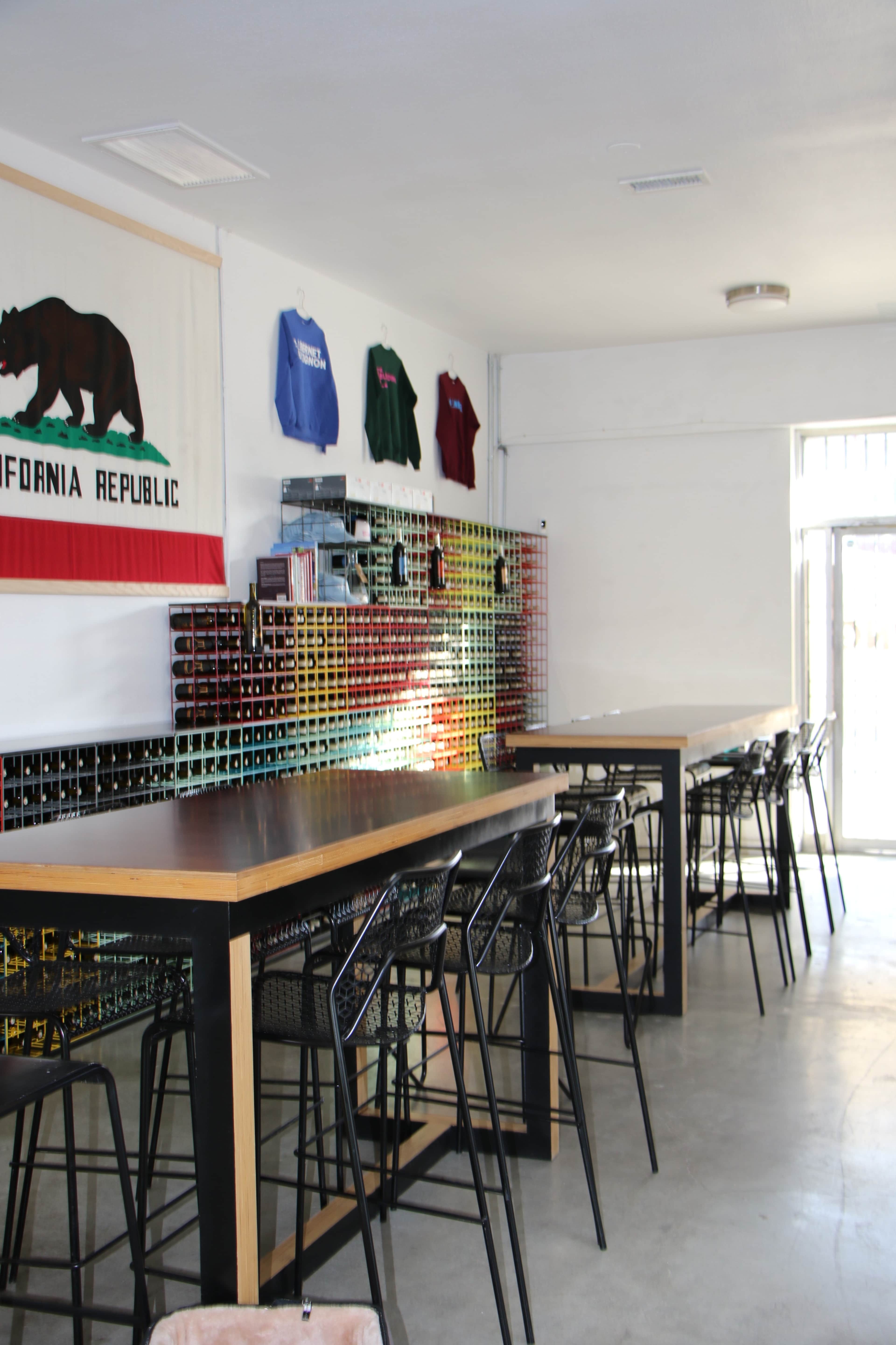 The image shows a spacious, minimalist indoor seating area featuring long tables and black chairs, decorated with a California Republic flag and colorful shirts on the wall.