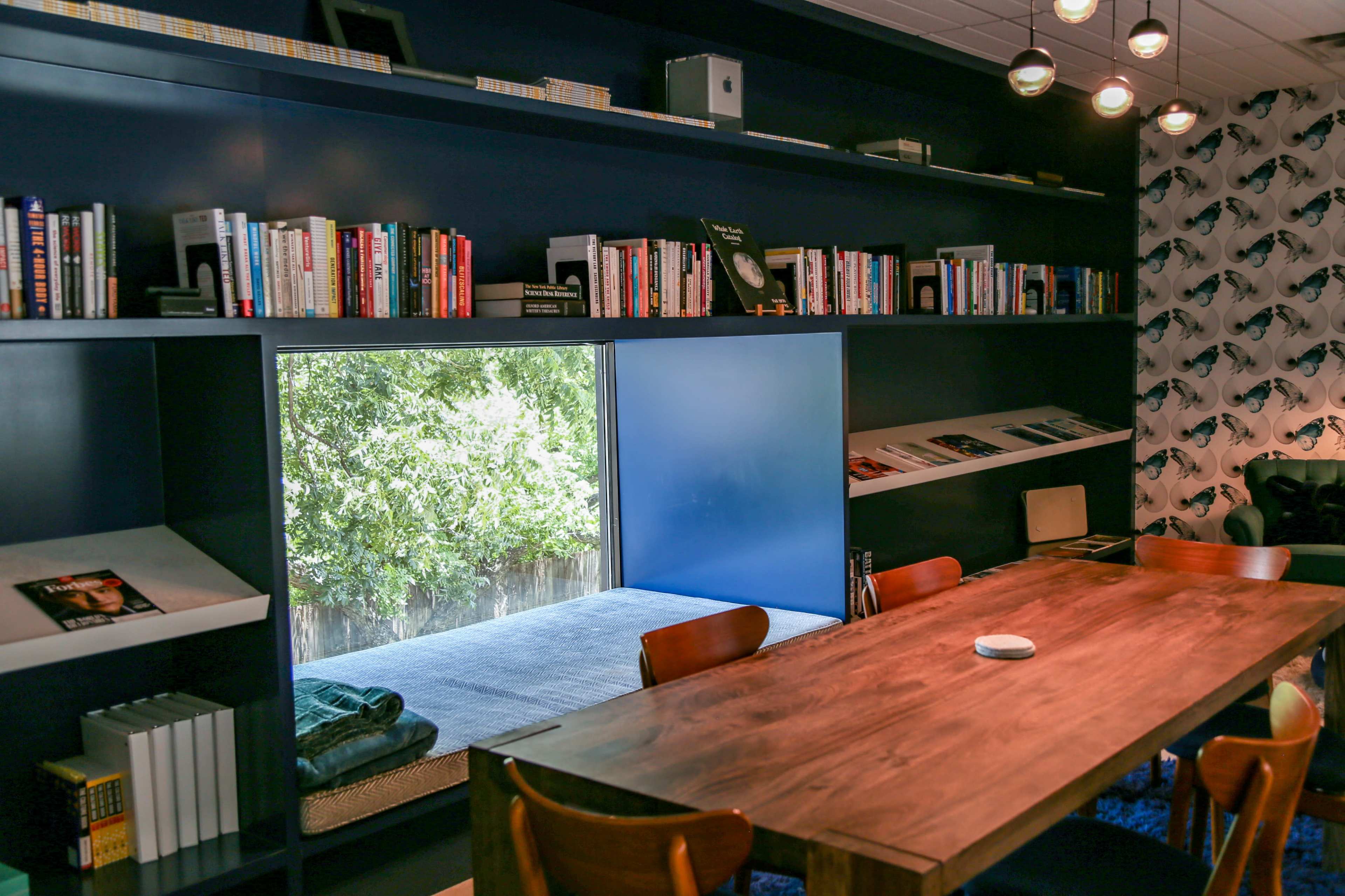 A spacious room features a long wooden table surrounded by chairs, with bookshelves filled with books lining the walls and a window nook offering a view of greenery outside.