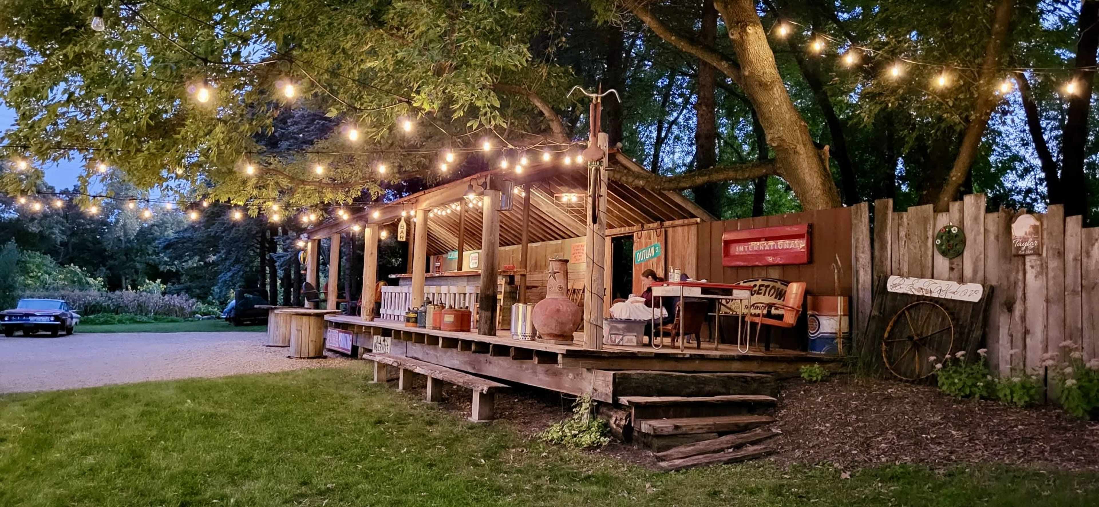 A wooden deck with string lights features tables and various items, surrounded by trees in a garden setting during twilight.