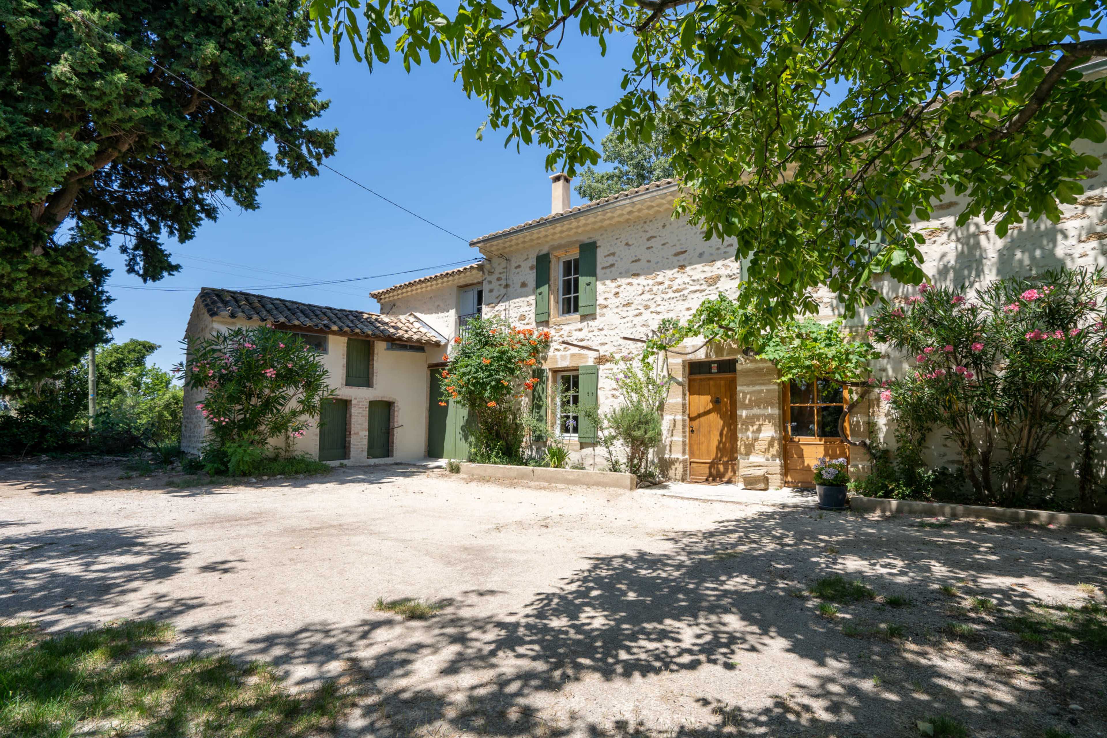 A two-story stone house with green shutters is surrounded by trees and shrubs in a gravel courtyard.